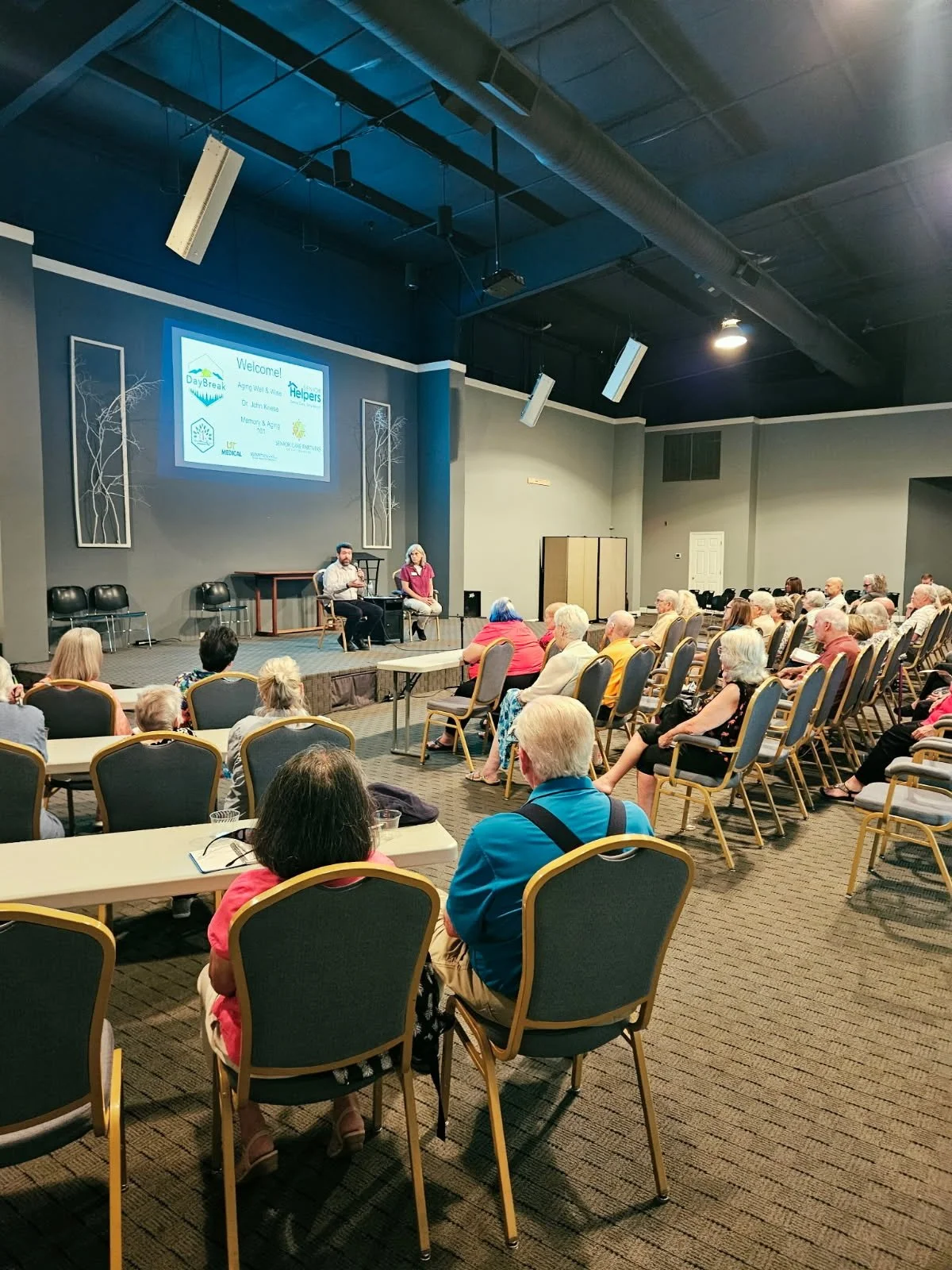 A group of elderly people seated in a conference room, listening to a panel discussion with two speakers at the front. The room has a large projection screen displaying a welcome slide.
