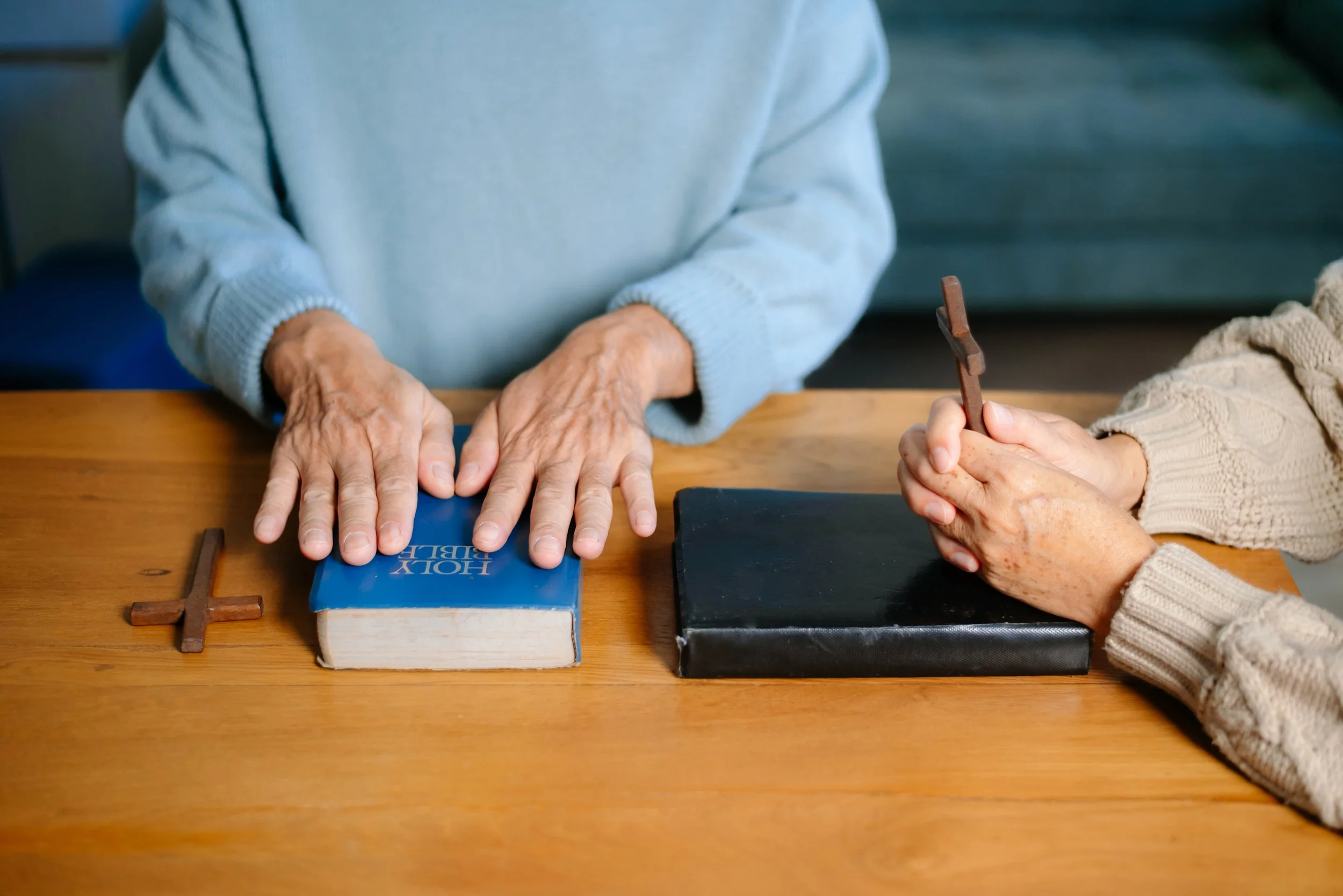 Two elderly women sitting at a wooden table with Christian symbols, including a small cross, and books, one woman holding a small cross and the other with her hands resting on a book.