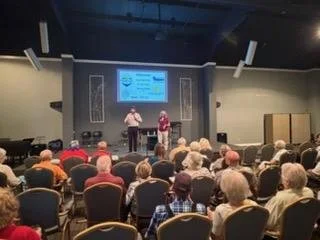 A speaker stands on stage in front of a projected screen, addressing an audience seated in rows at an indoor event or conference.