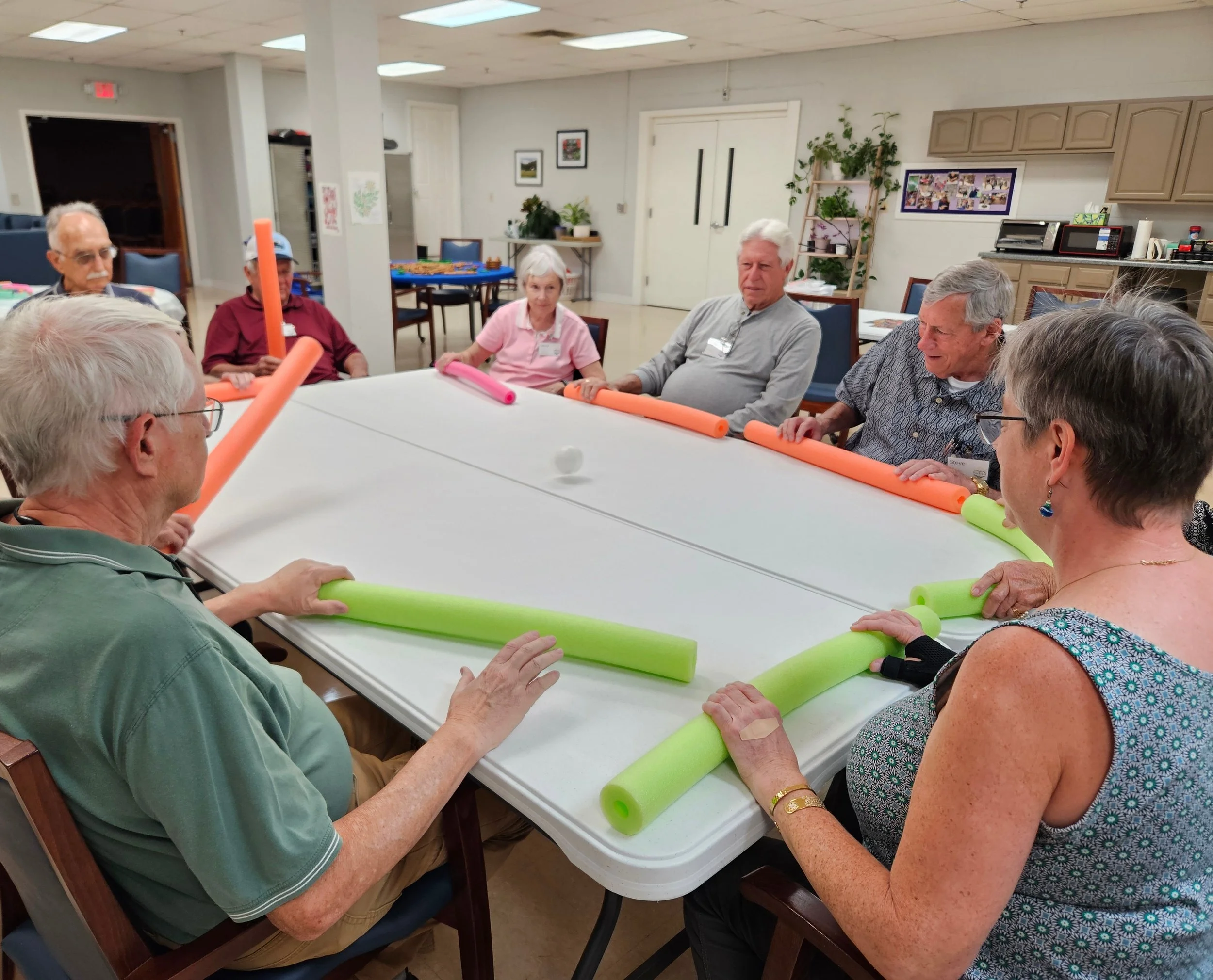 Group of elderly people participating in a seated exercise session using pool noodles, sitting around a white table in a community center.