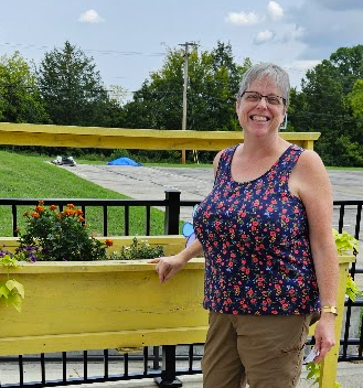 A smiling woman standing outside near a yellow planter box with flowers, with a black railing and green trees in the background.