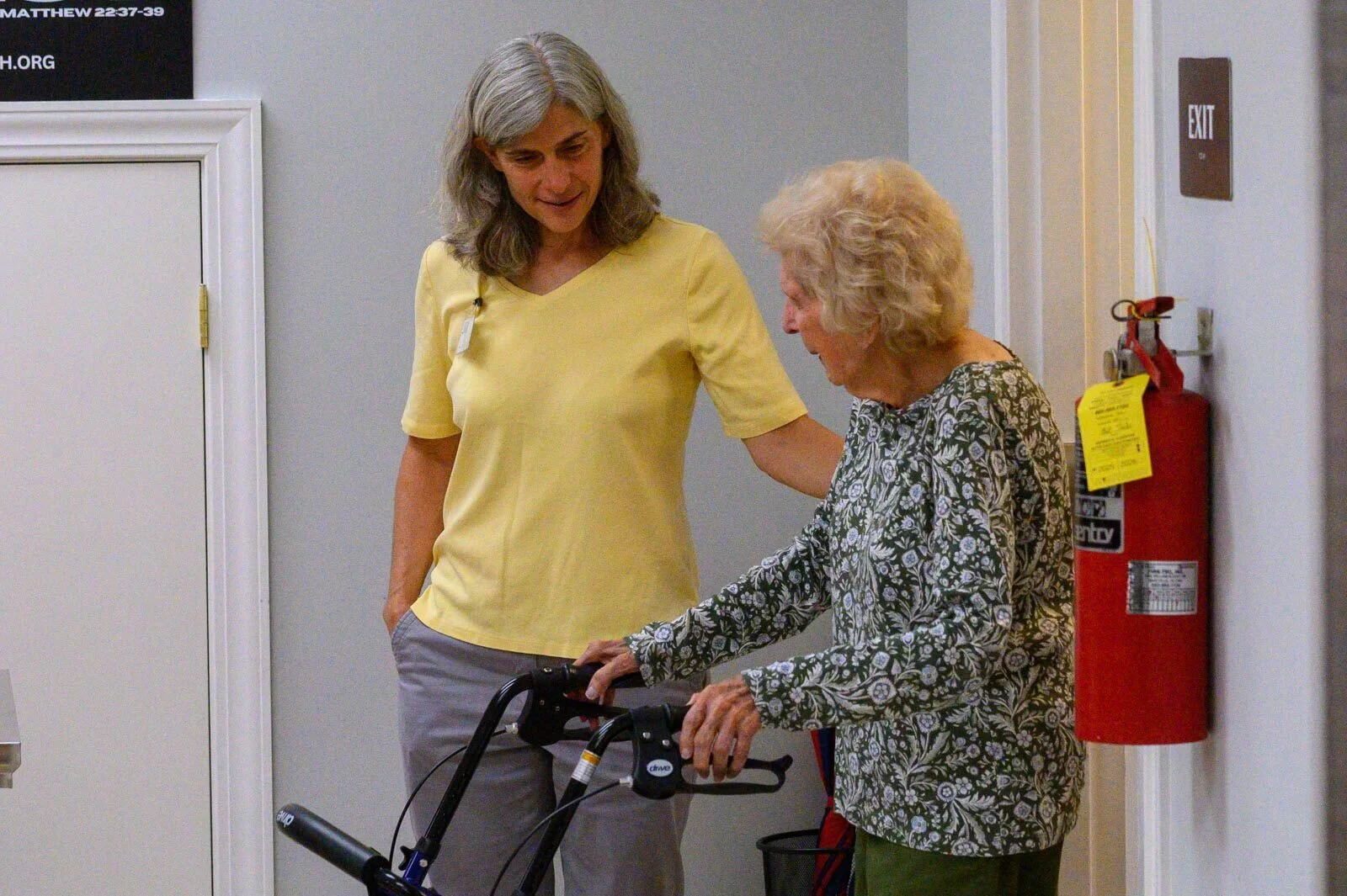 A young woman with gray hair and a yellow shirt assisting an elderly woman with curly gray hair, who is using a walker, in a hallway with a fire extinguisher on the wall.