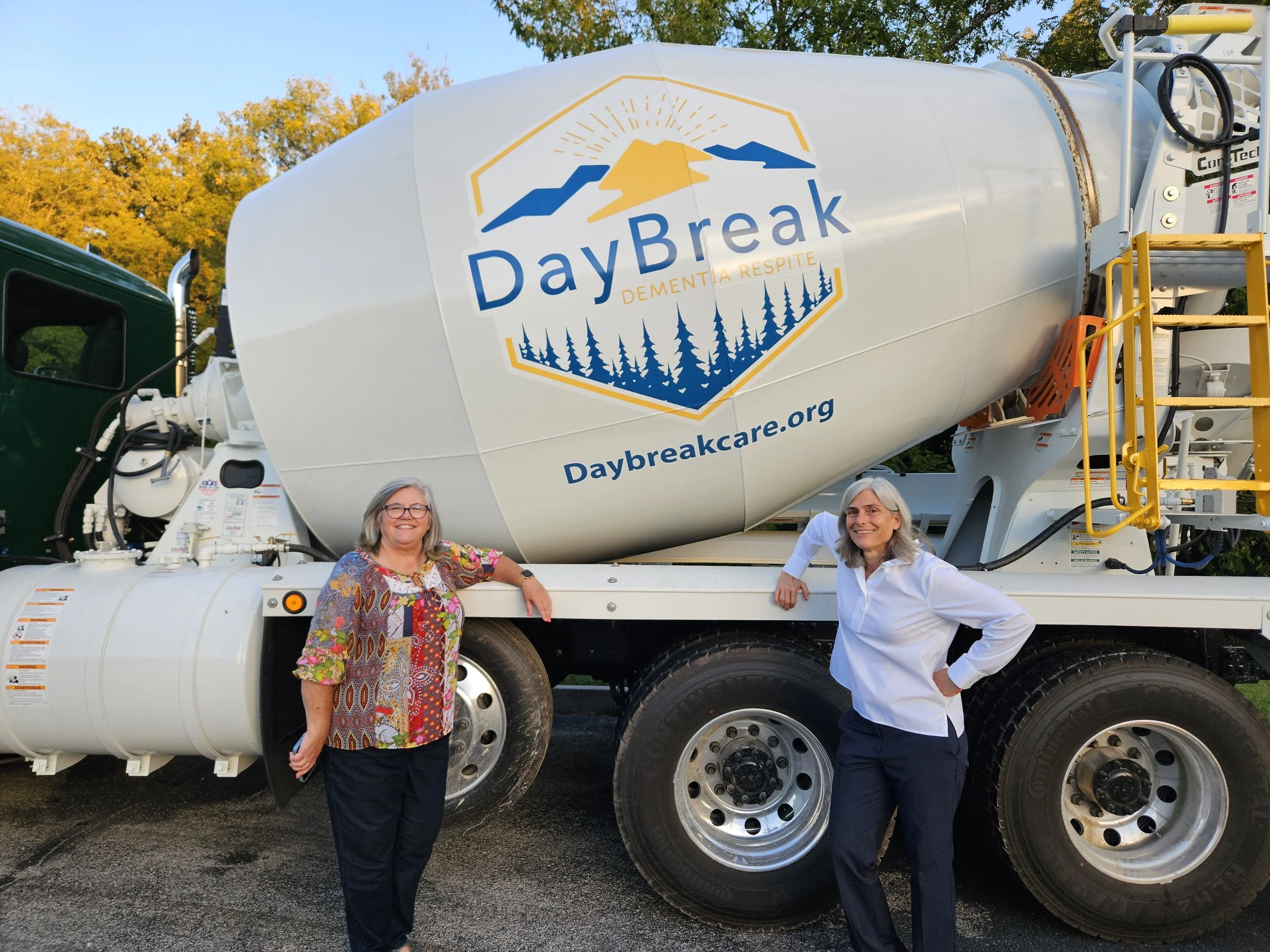 Two women standing next to a large vehicle with a mural of a mountain and sunset, reading "DayBreak Dementia Respite" and a website URL. The women are smiling and dressed casually, posing outdoors with trees in the background.