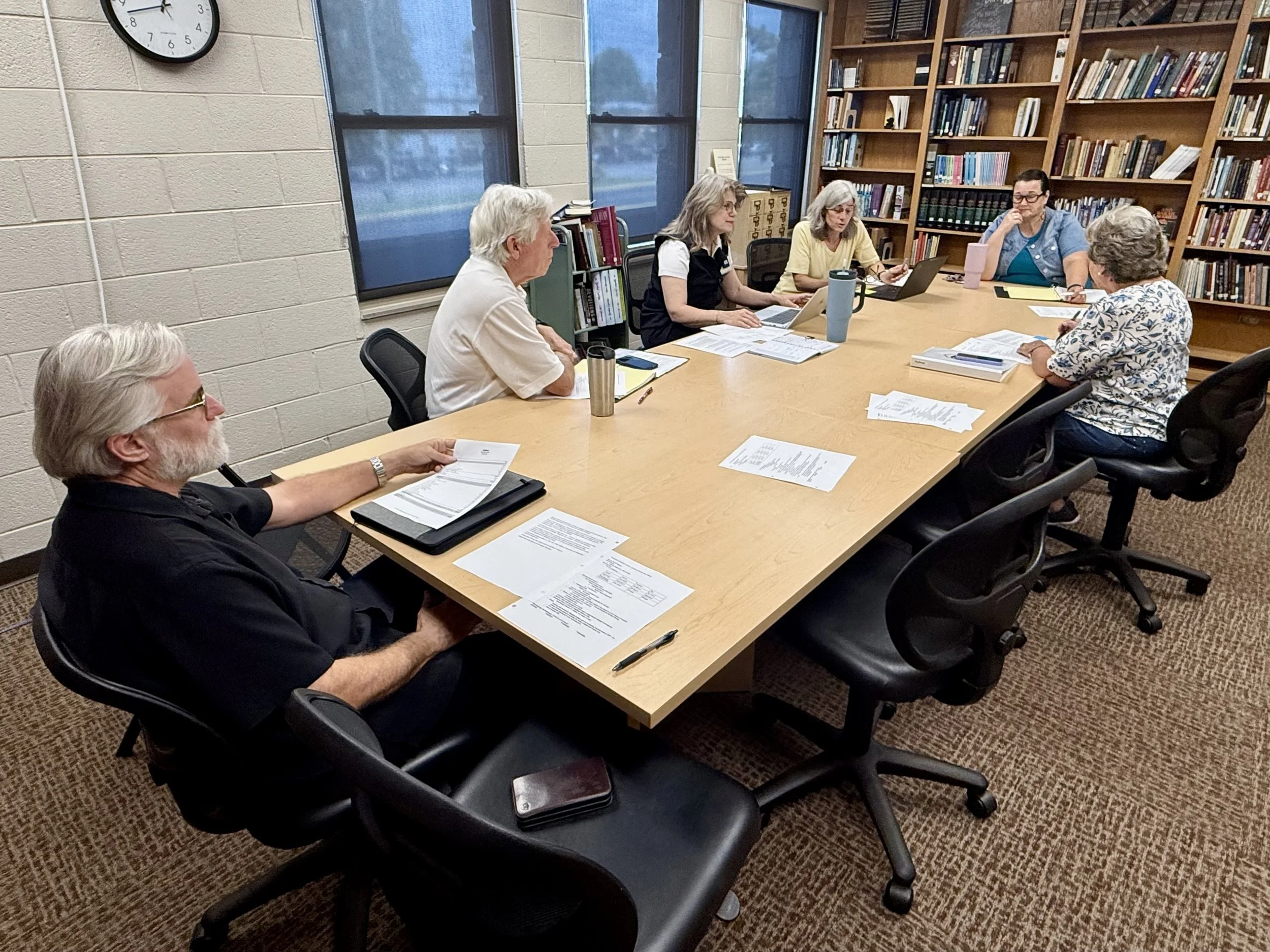 A group of six people sitting around a rectangular table in a library, engaged in discussion with notebooks, papers, and laptops, with bookshelves and windows in the background.