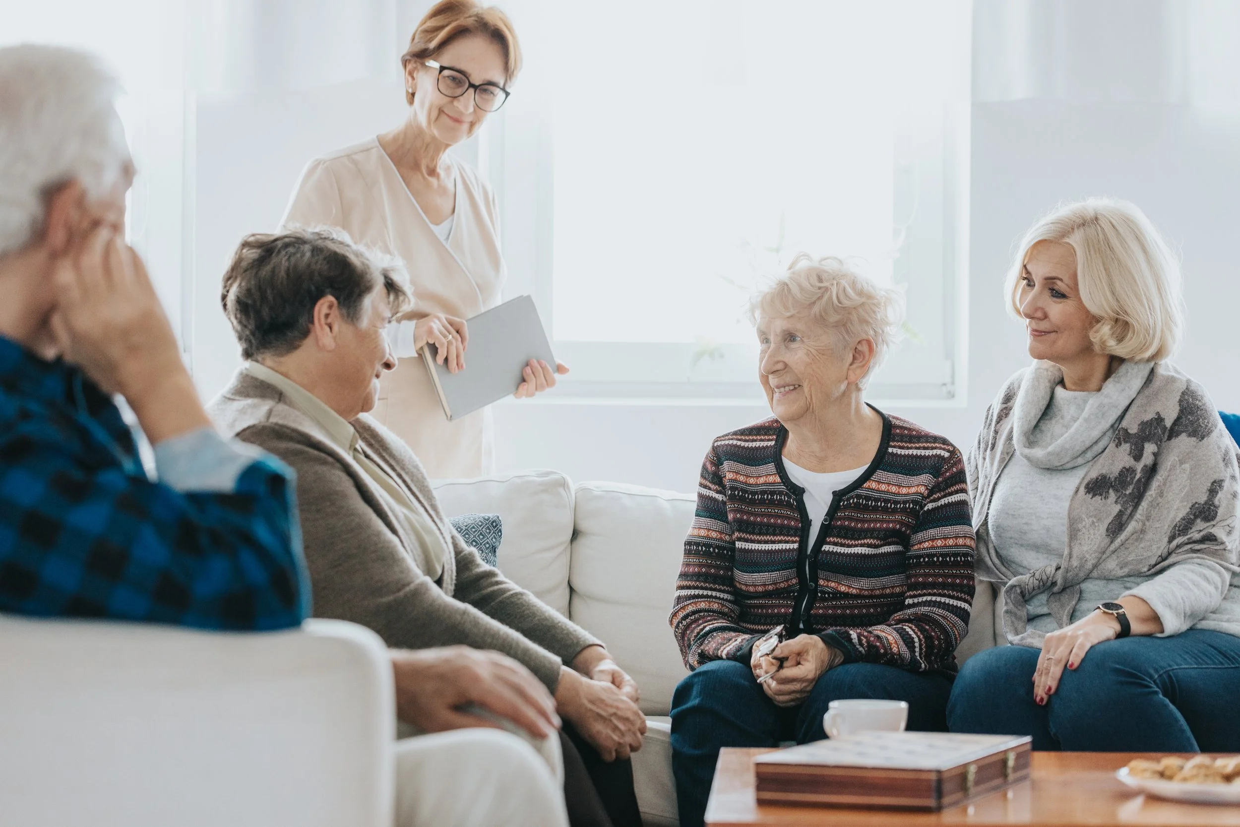 A woman standing and holding a folder, talking to a group of older adults sitting in a living room.