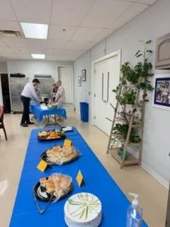A table with various desserts, including a cake with a green decoration, a pie, and a plate of cookies, set up in a room with people serving food in the background.
