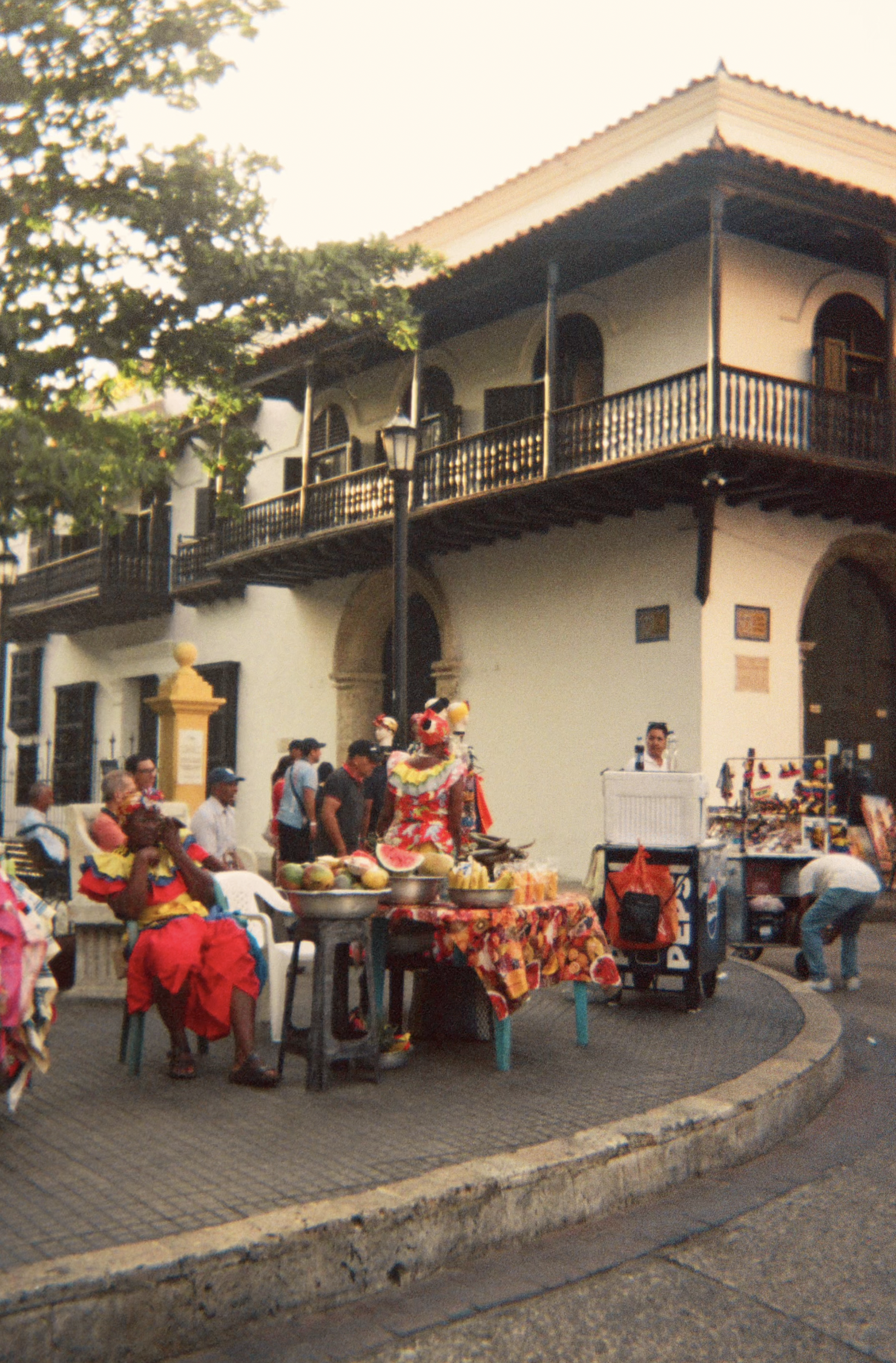 Street scene with vendors and shoppers, colorful costumes, and a historic building with a balcony in the background.