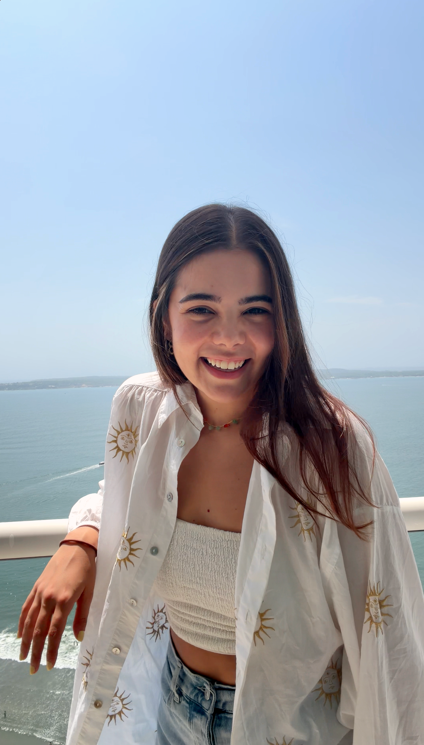 A young woman smiling outdoors near the water, wearing a white button-up shirt with sun and moon designs over a white top and denim shorts.