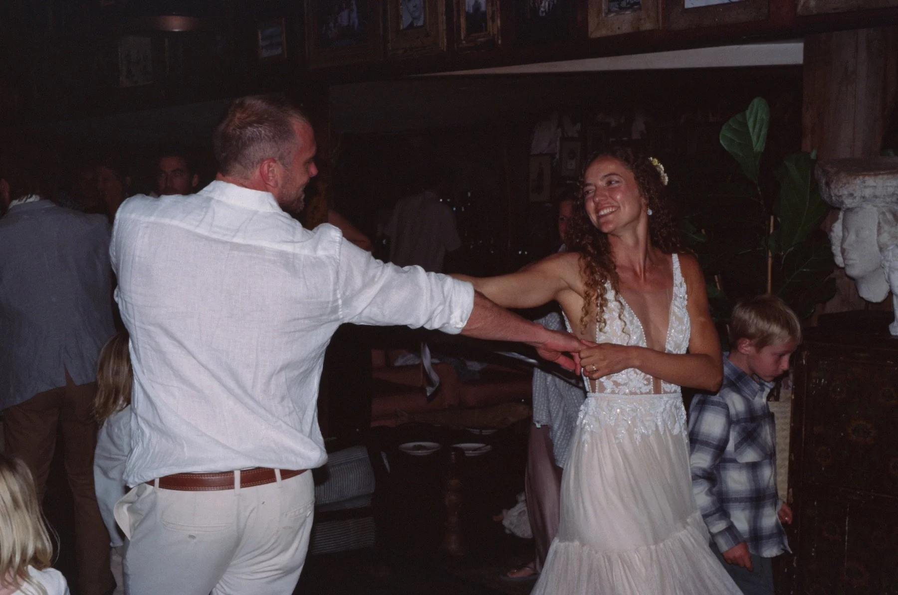 A man and woman are dancing together, holding hands, at a lively indoor event. The woman is smiling and wearing a white dress with lace details, while the man is dressed in a white shirt with rolled-up sleeves. Other people are in the background, som