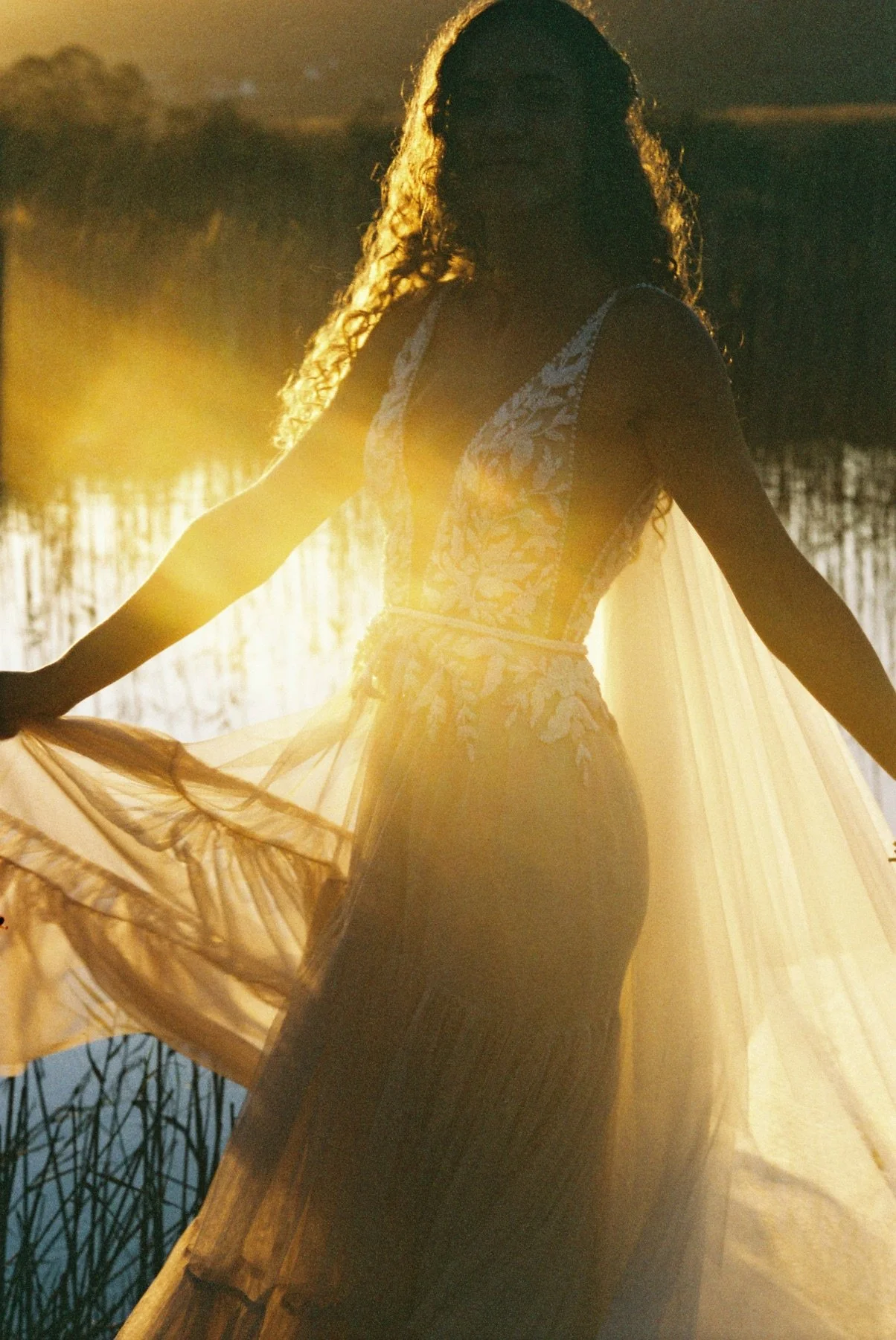A woman with curly hair wearing a light-colored, lace dress holding a flowing fabric, standing near a body of water during sunset.