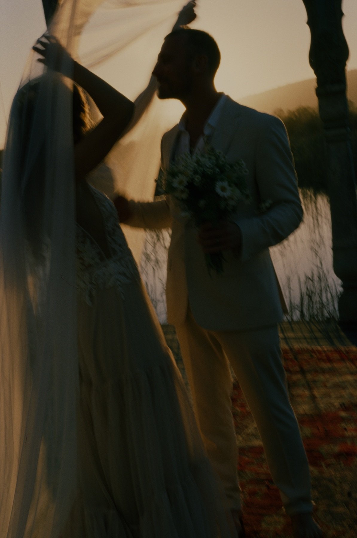 Silhouette of a man and woman with wedding attire exchanging vows outdoors at sunset, holding a bouquet of flowers.