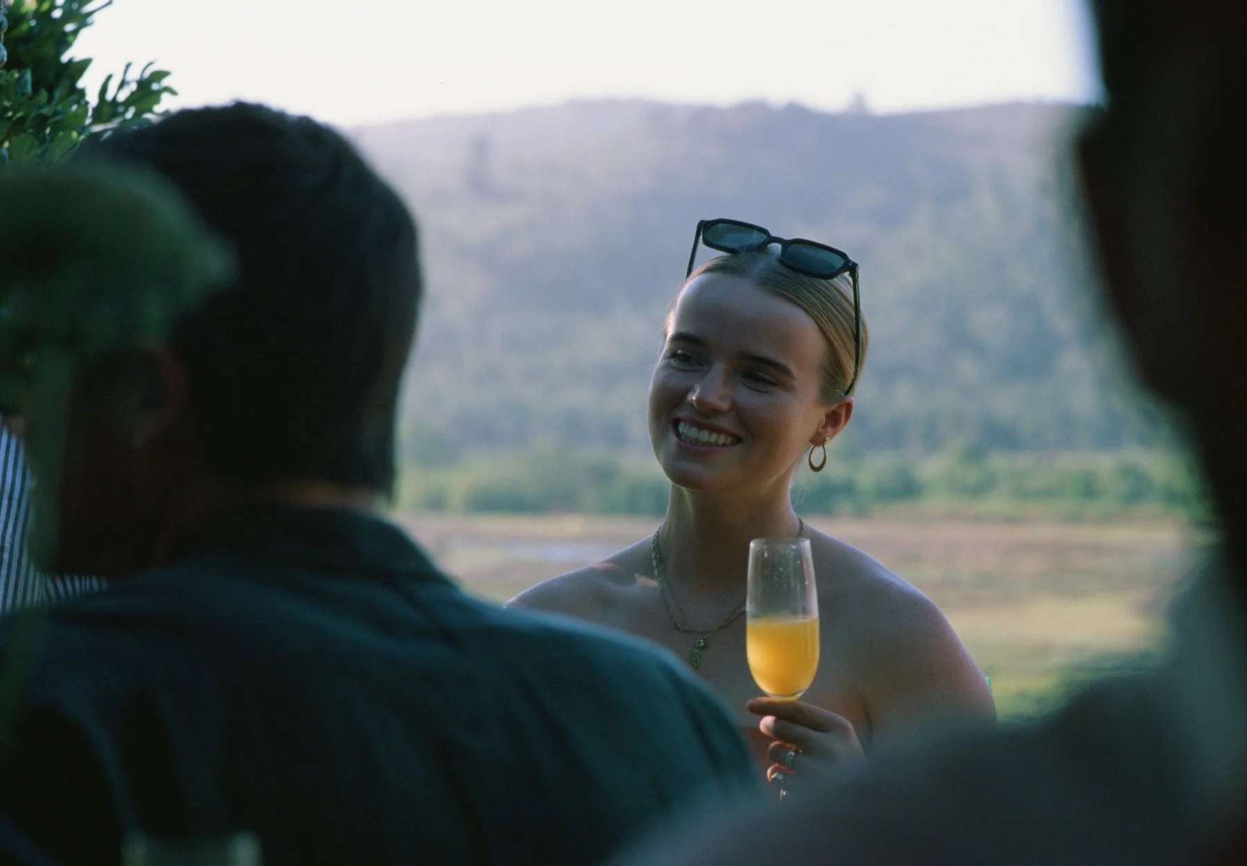 A woman holding a glass of orange drink, smiling, wearing sunglasses on her head, in an outdoor setting with hills in the background.