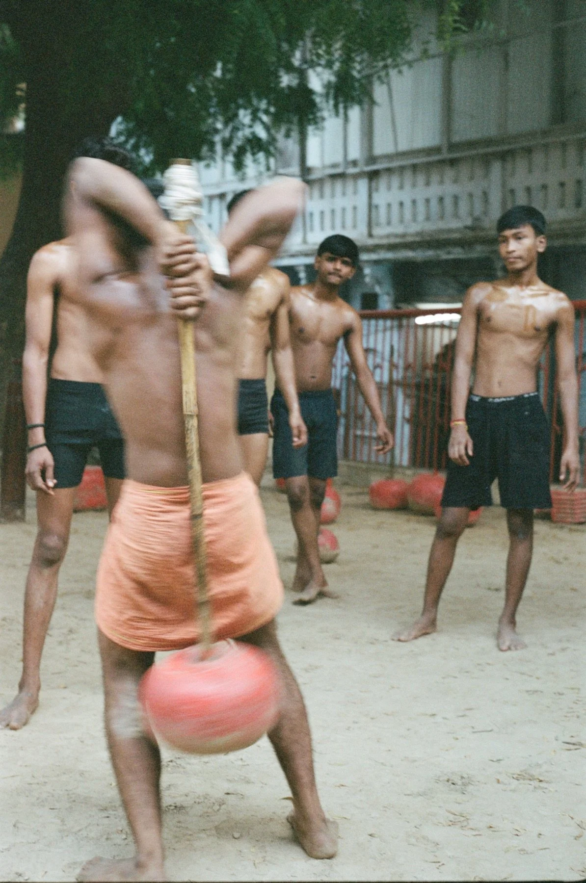 A group of young boys, shirtless and wearing shorts, playing a game with a ball and a stick outdoors on a sandy ground. Some boys are watching while others are actively participating.