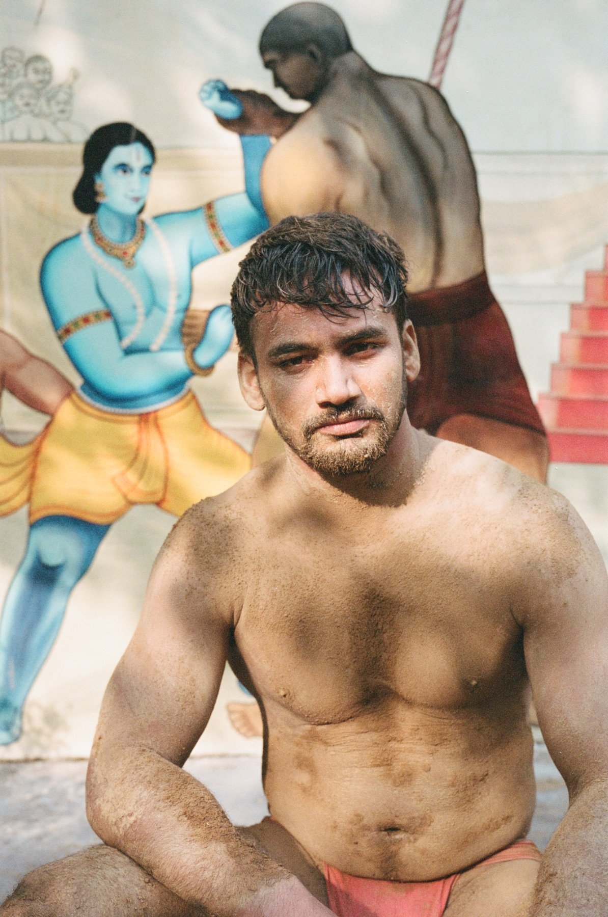 A shirtless young man with a beard and messy hair sitting in front of a painted wall featuring characters from Hindu mythology, including Lord Krishna with blue skin, wearing traditional gold and orange clothing and jewelry, and a muscular figure wit