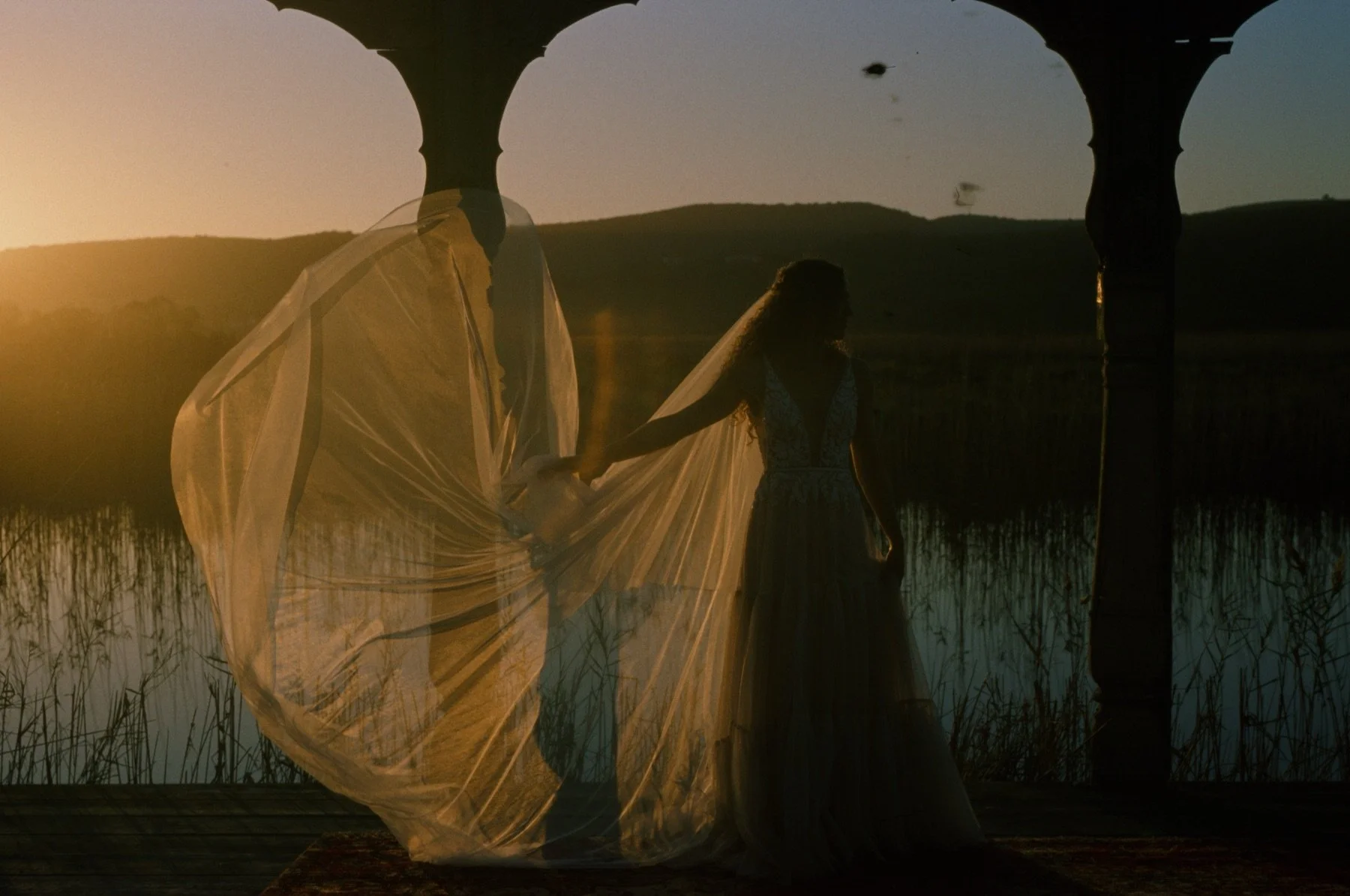 Two women holding hands, blurred silhouette, wearing flowing dresses, standing in a marsh at sunset with water and reeds, silhouetted against the sky, in a scenic outdoor setting.