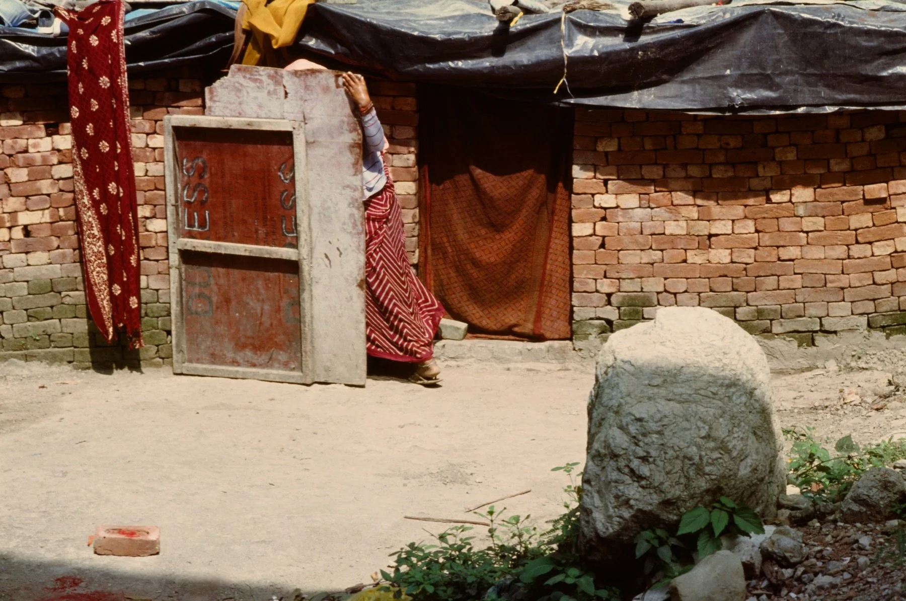 A woman in traditional clothing is standing outside a brick building, holding a sign or board in front of her face. The building has a dark tarp roof, and there are various fabrics hanging on the wall. The ground is sandy, and a large rock and some s