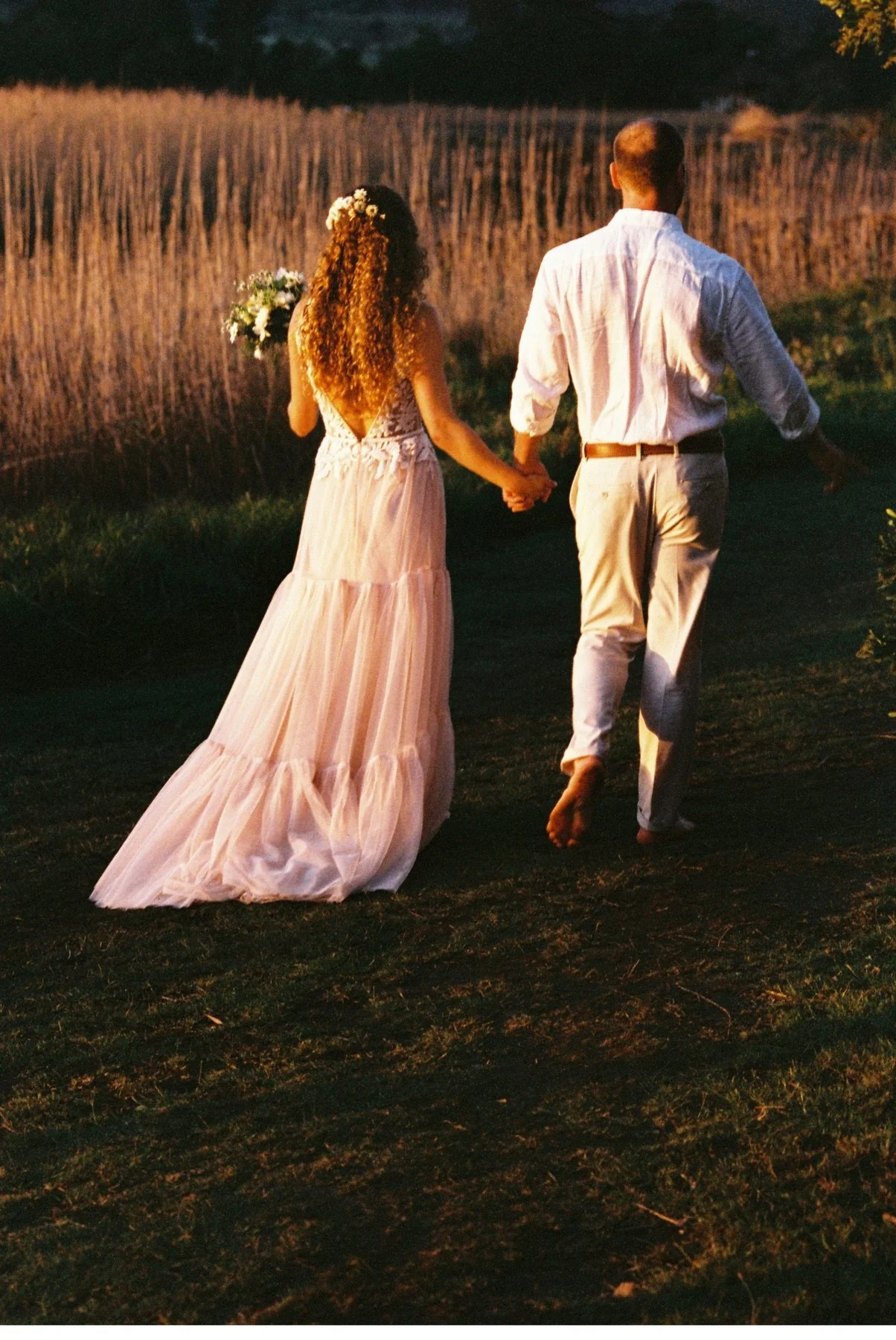 A bride and groom walking barefoot hand-in-hand through a grassy area at sunset, with the bride holding a bouquet of flowers.
