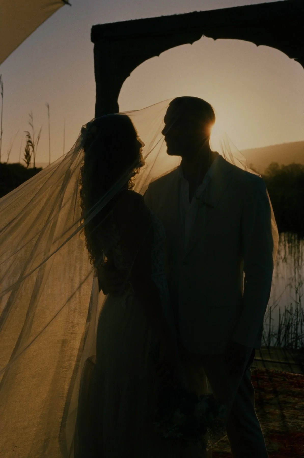 Silhouetted bride and groom standing close together outdoors during sunset, with a veil lightly covering her face and a stone arch in the background.