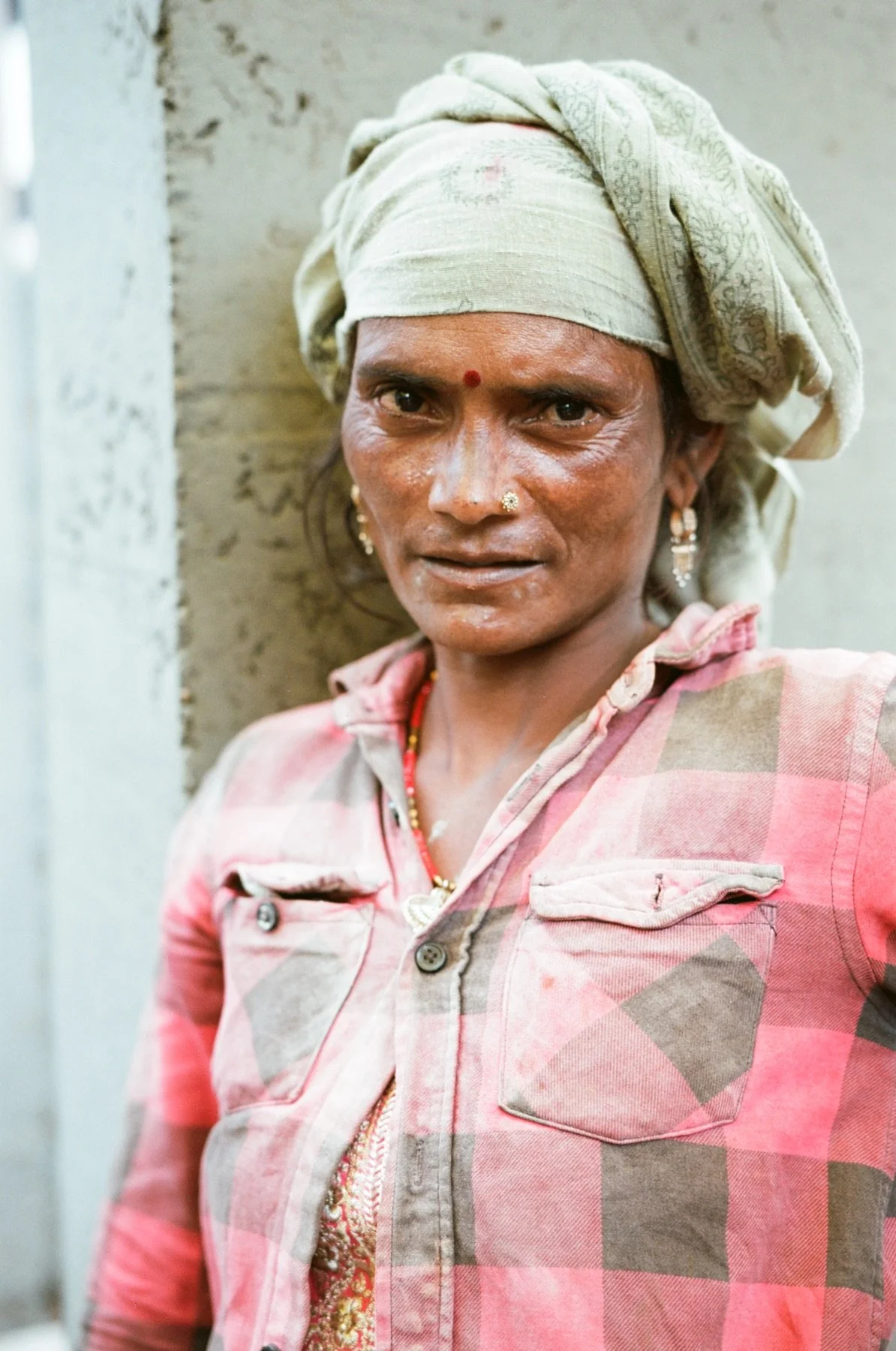 A woman with dark skin wearing a beige headscarf, earrings, and a pink checkered shirt with a slight stain leaning against a concrete wall.