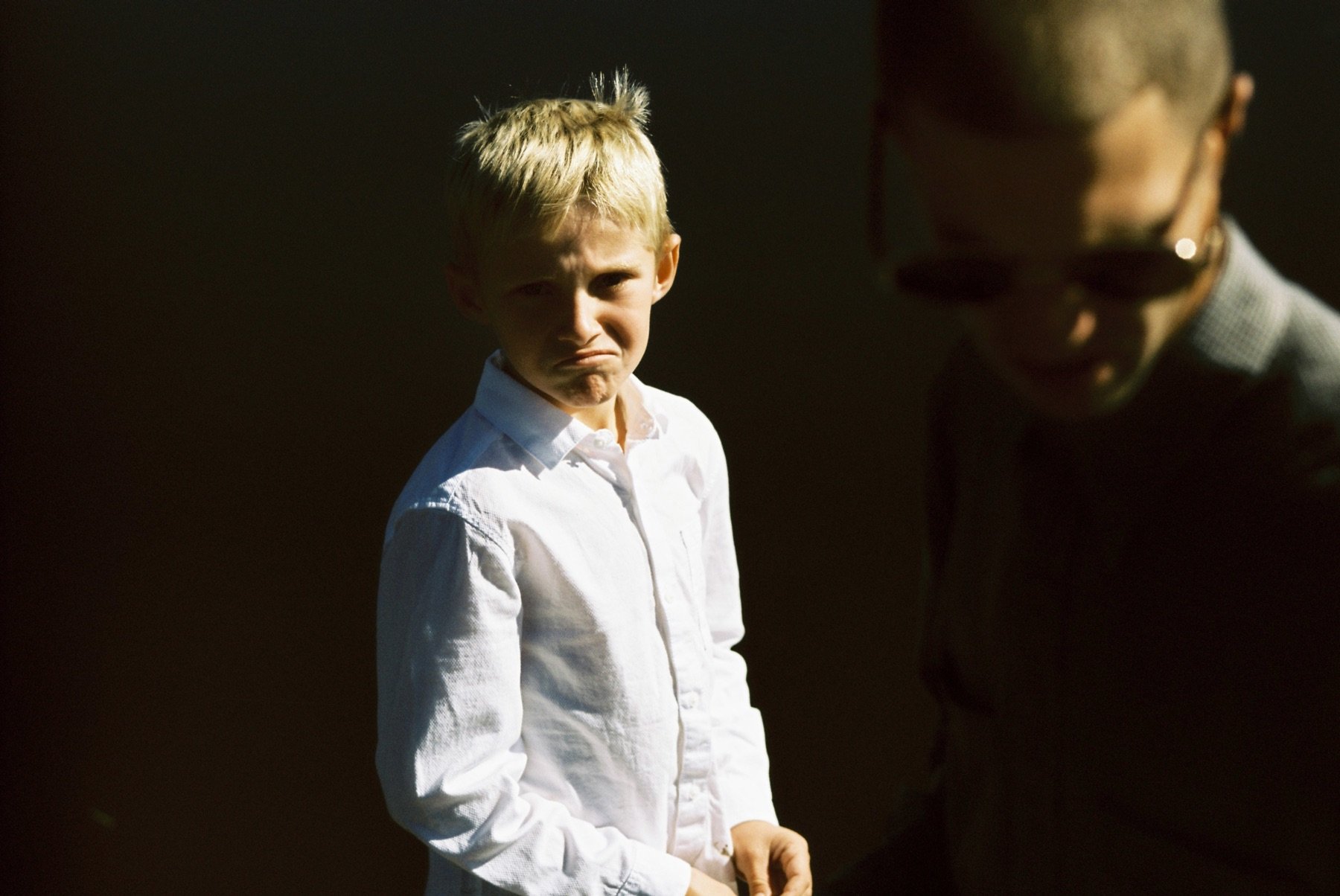 A boy with a sad or disappointed expression stands in shadow, wearing a white shirt, while a person with glasses and a checkered shirt looks down in the dark.