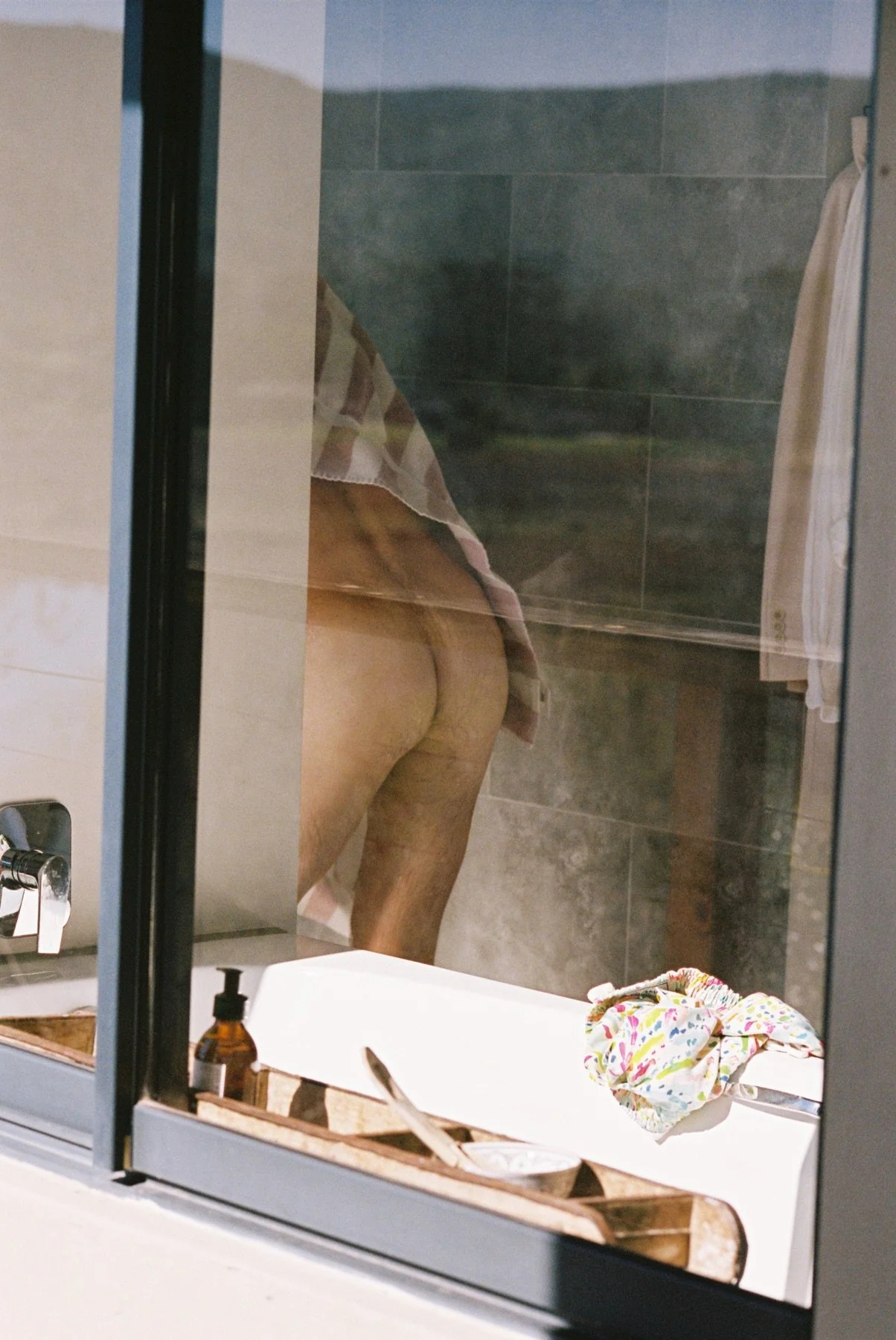 A woman taking a mirror selfie in a bathroom, showing her lower back and buttocks. The bathroom has gray tiled walls, a white sink with a colorful cloth and a soap dispenser on it, and a mirror reflecting the scene.
