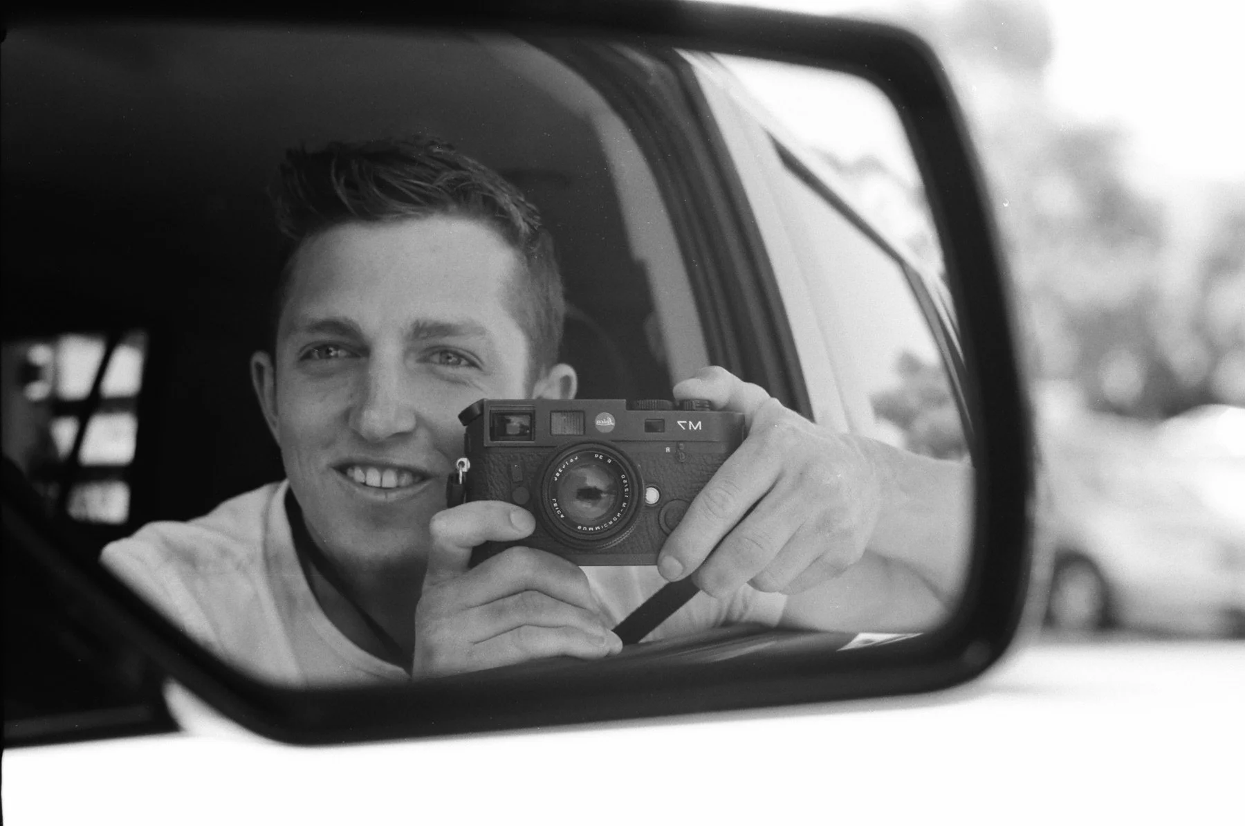 A young man taking a selfie with a camera in his hand, smiling, reflected in a car's side mirror.