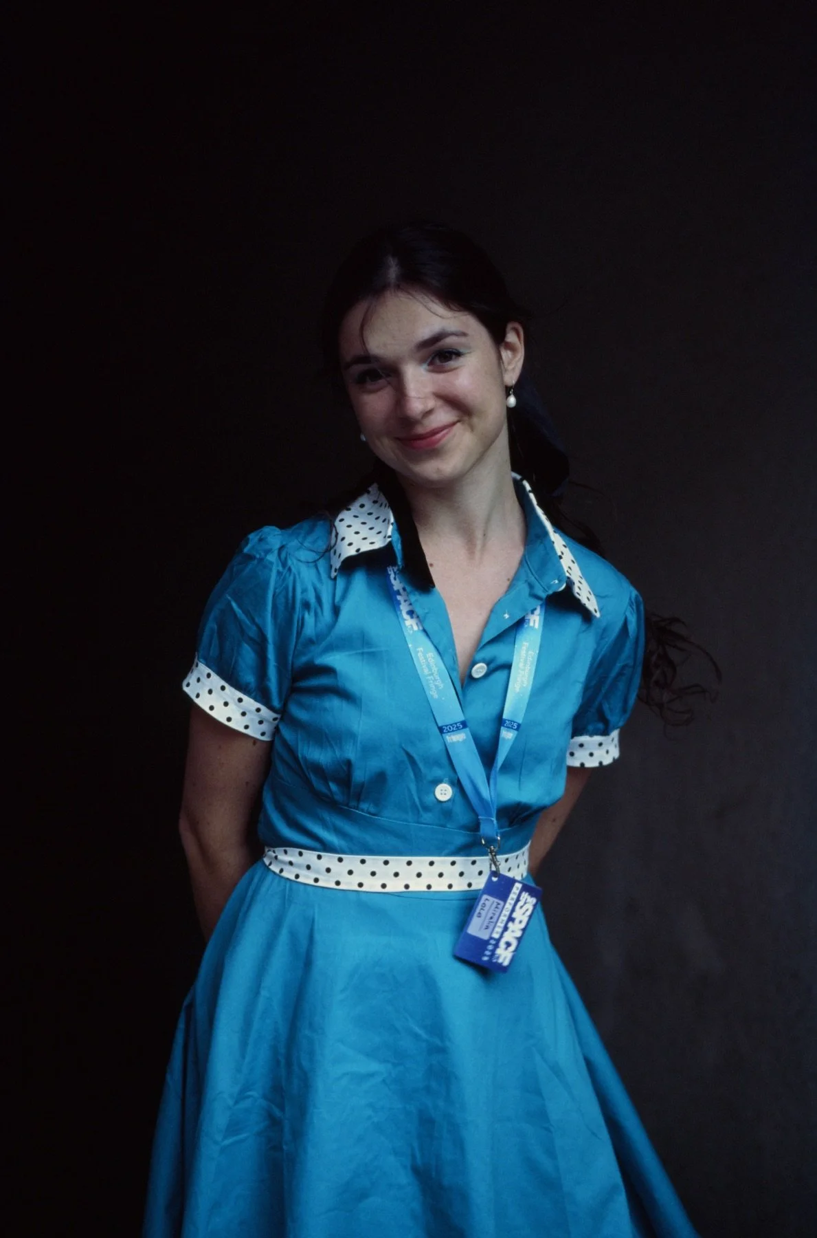 A young woman with dark hair in a retro blue dress with polka dot accents, smiling with a lanyard around her neck, standing against a plain dark background.