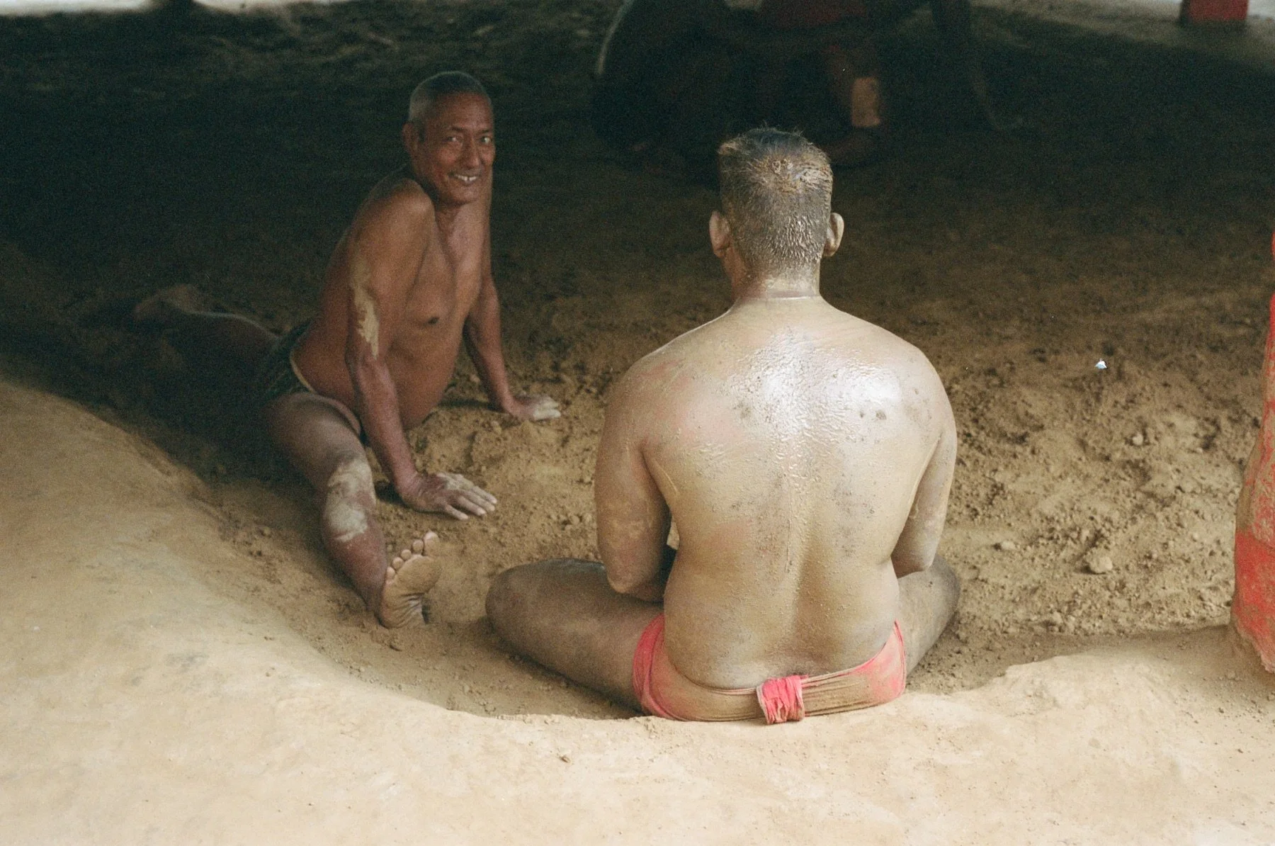 Two shirtless men sitting on the ground in a dark, earthy setting, possibly a cave or enclosed space, with one man facing away from the camera and the other looking towards the camera smiling.