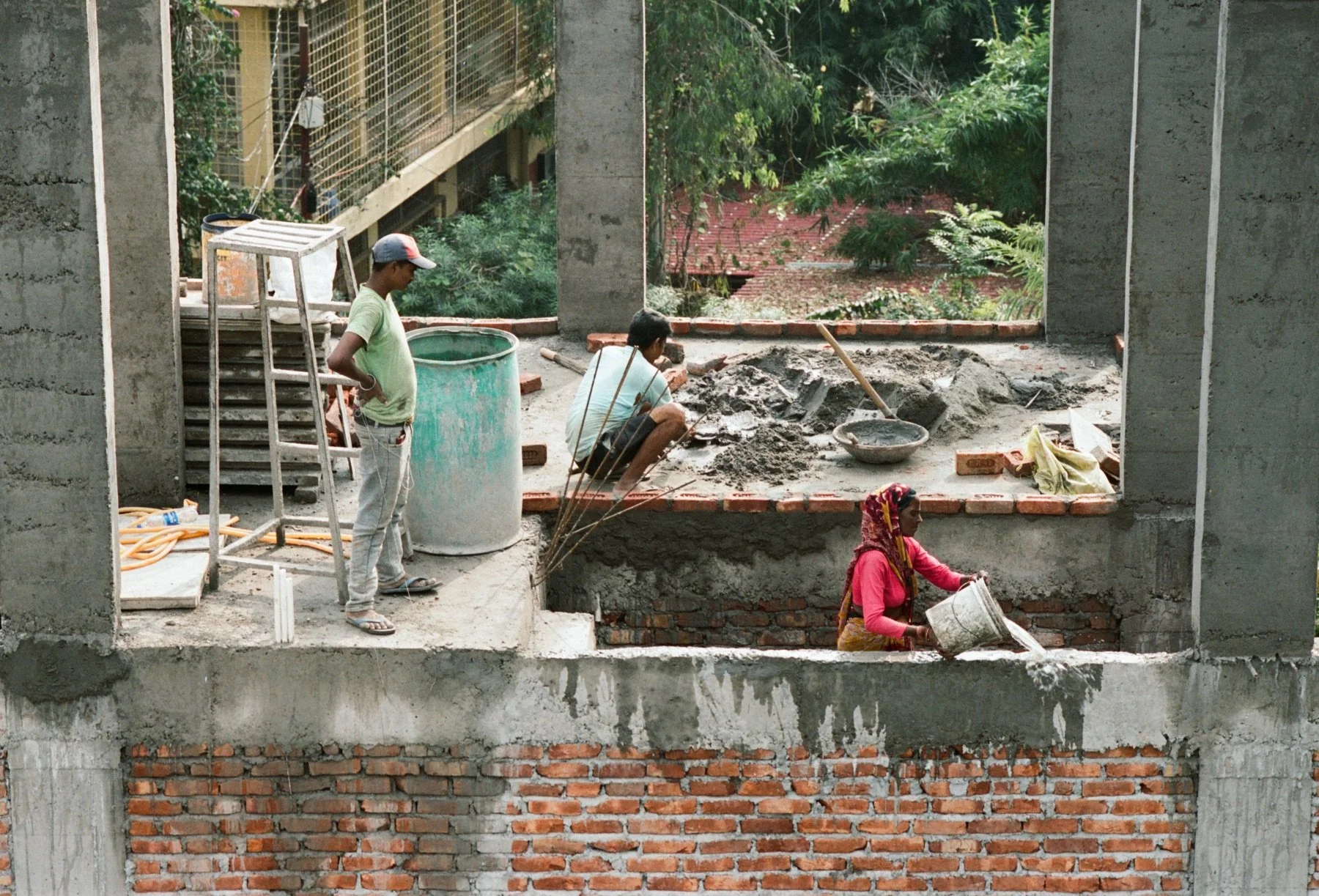 People working on construction of a building, with one person sitting and another pouring concrete, while a woman stands nearby holding a bucket. The building's brick and concrete structure is visible, with open spaces for windows.