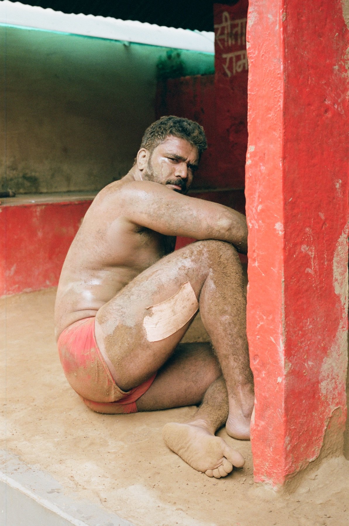 A shirtless man with sand on his skin and mud on his body, sitting on the ground with his knees pulled up to his chest, leaning against a red wall, wearing red shorts, in a sandy environment.