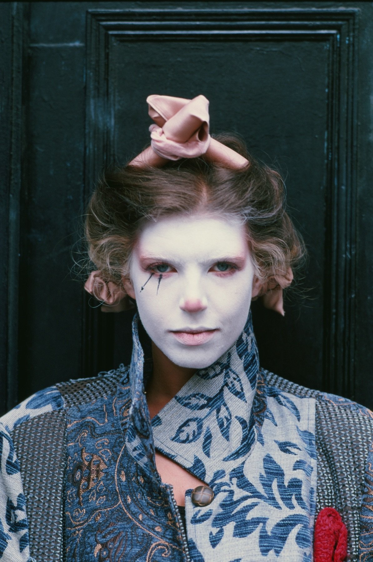 A person with pale face makeup, painted red nose, and dramatic eye makeup with black lines, wearing a patterned shirt and standing against a dark background.