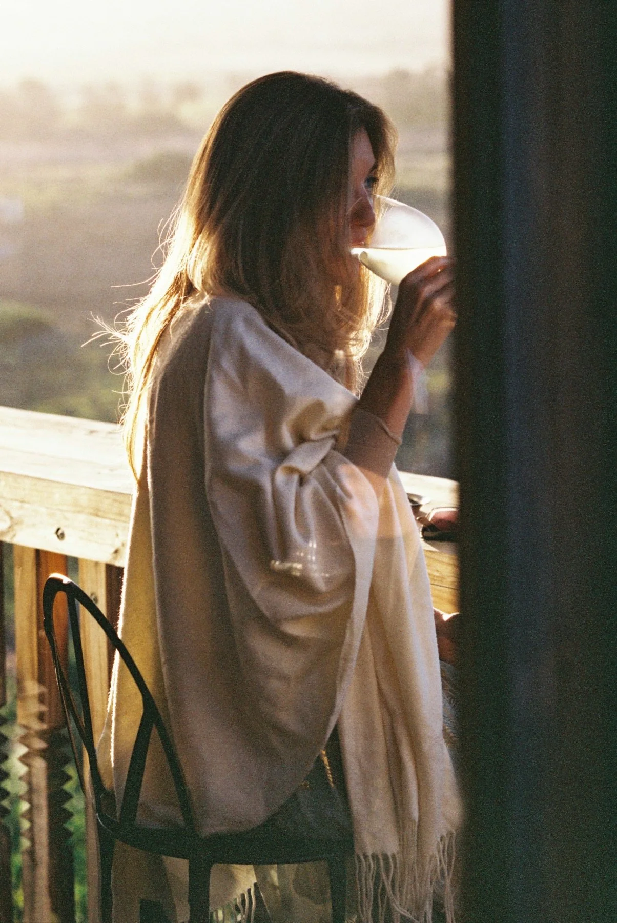 A woman with light brown hair drinking a cocktail on a wooden deck during sunset.