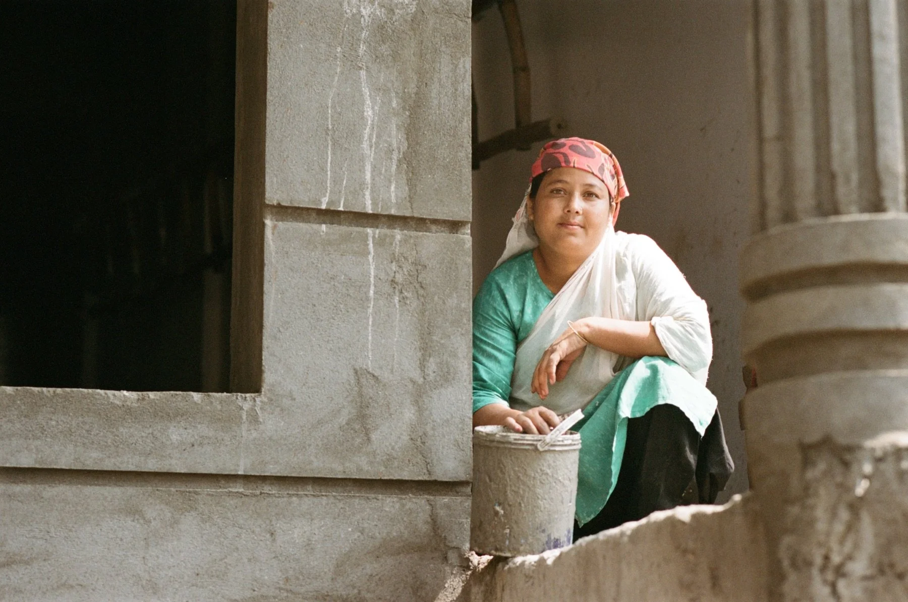 A woman wearing a colorful headscarf and a green and white traditional outfit sitting on a ledge inside a building with unfinished concrete walls.