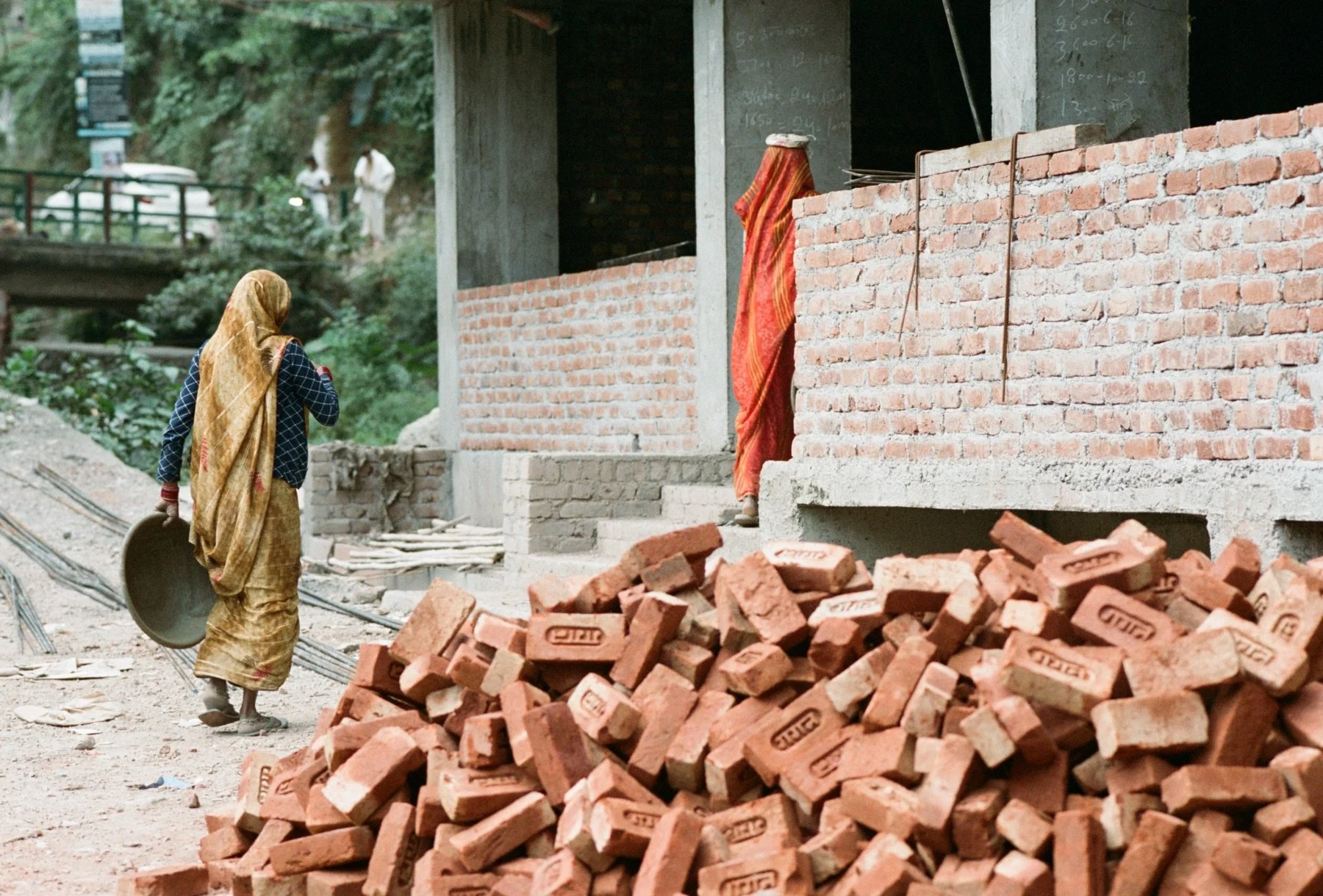 A woman carrying a metal bowl walking past a pile of red bricks at a construction site with a partially built brick wall. In the background, a person is seen entering a building, and trees are visible beyond the construction area.