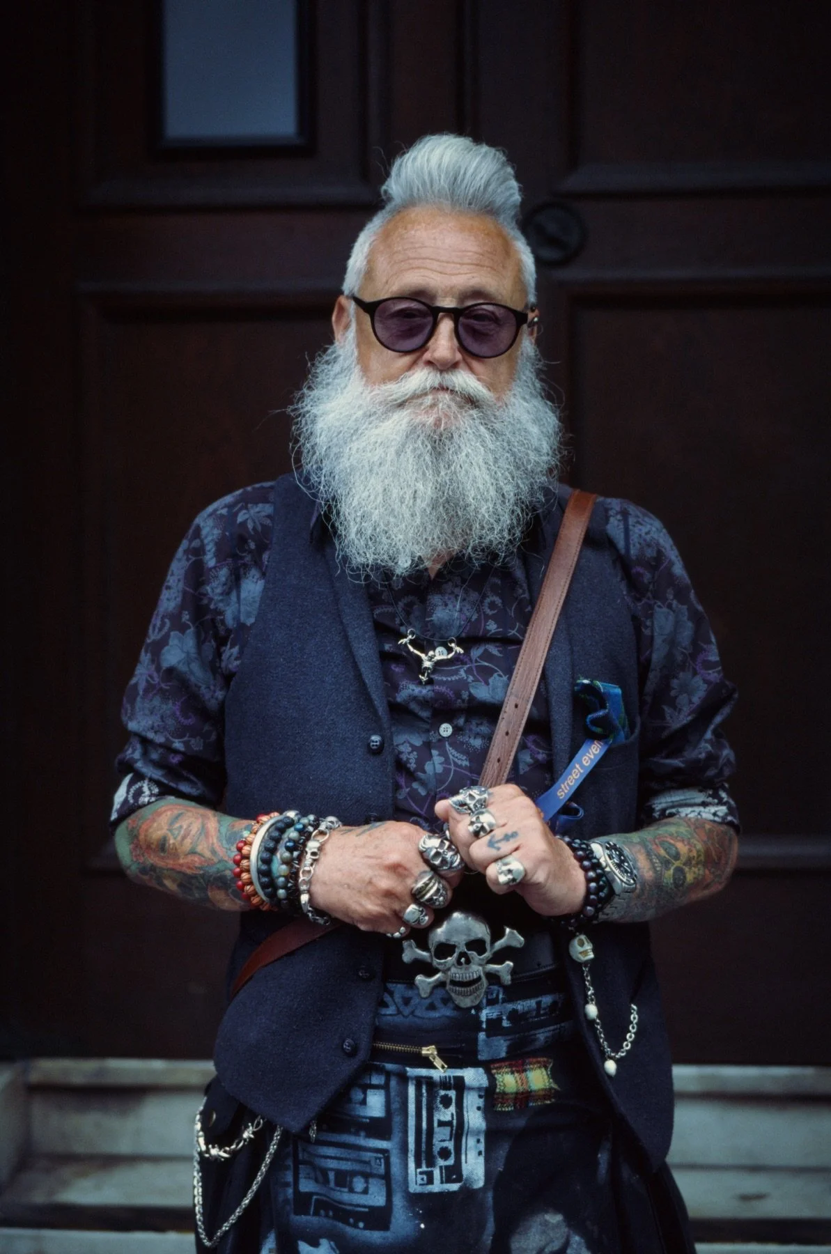 An older man with a long white beard and gray hair, wearing black sunglasses and a dark floral shirt, standing in front of a wooden background. He has tattoos on his arms and is wearing various rings, bracelets, and necklaces, including a prominent s