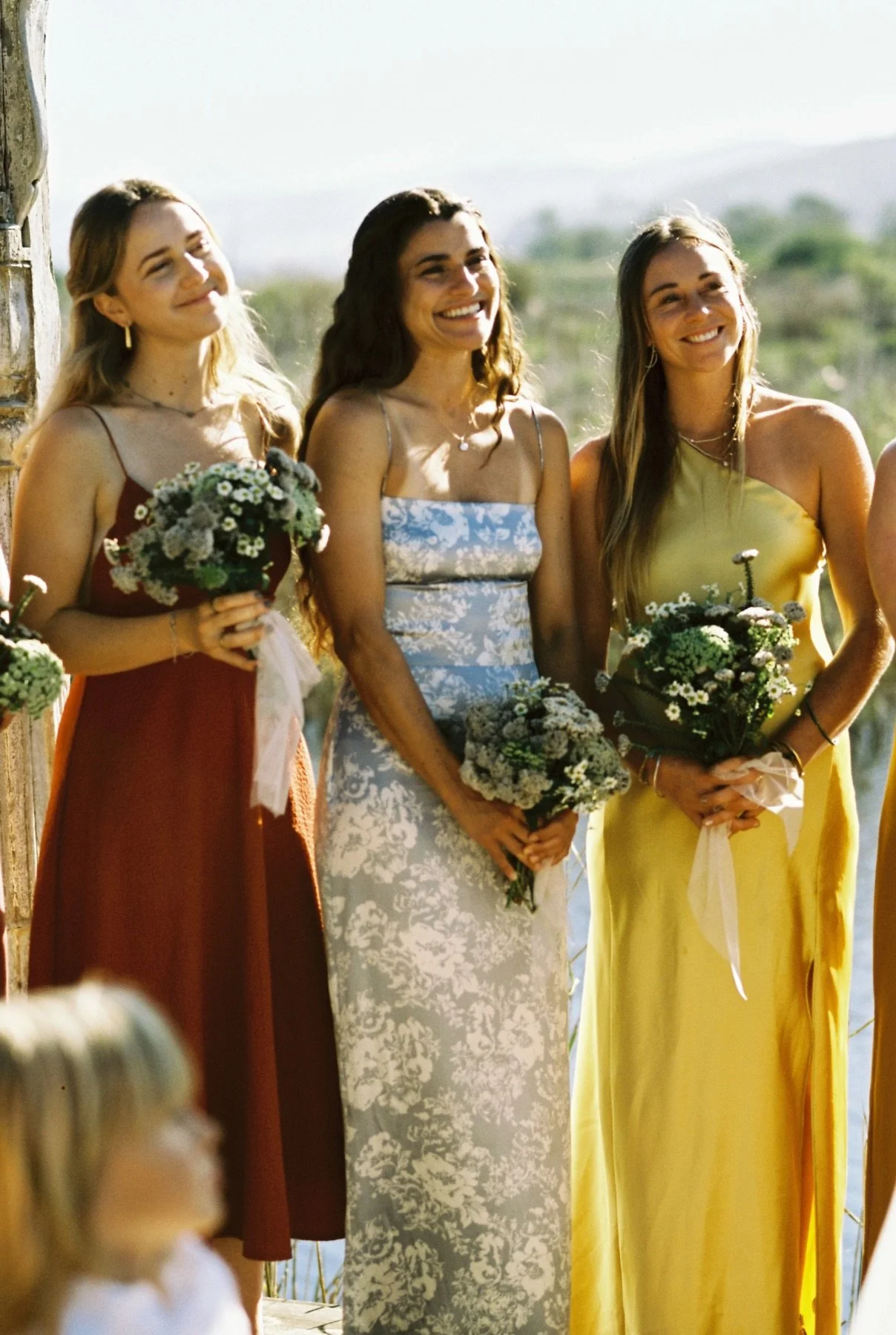 Three women smiling, holding bouquets of flowers, standing outdoors during a celebration, with a scenic background of hills and sky.