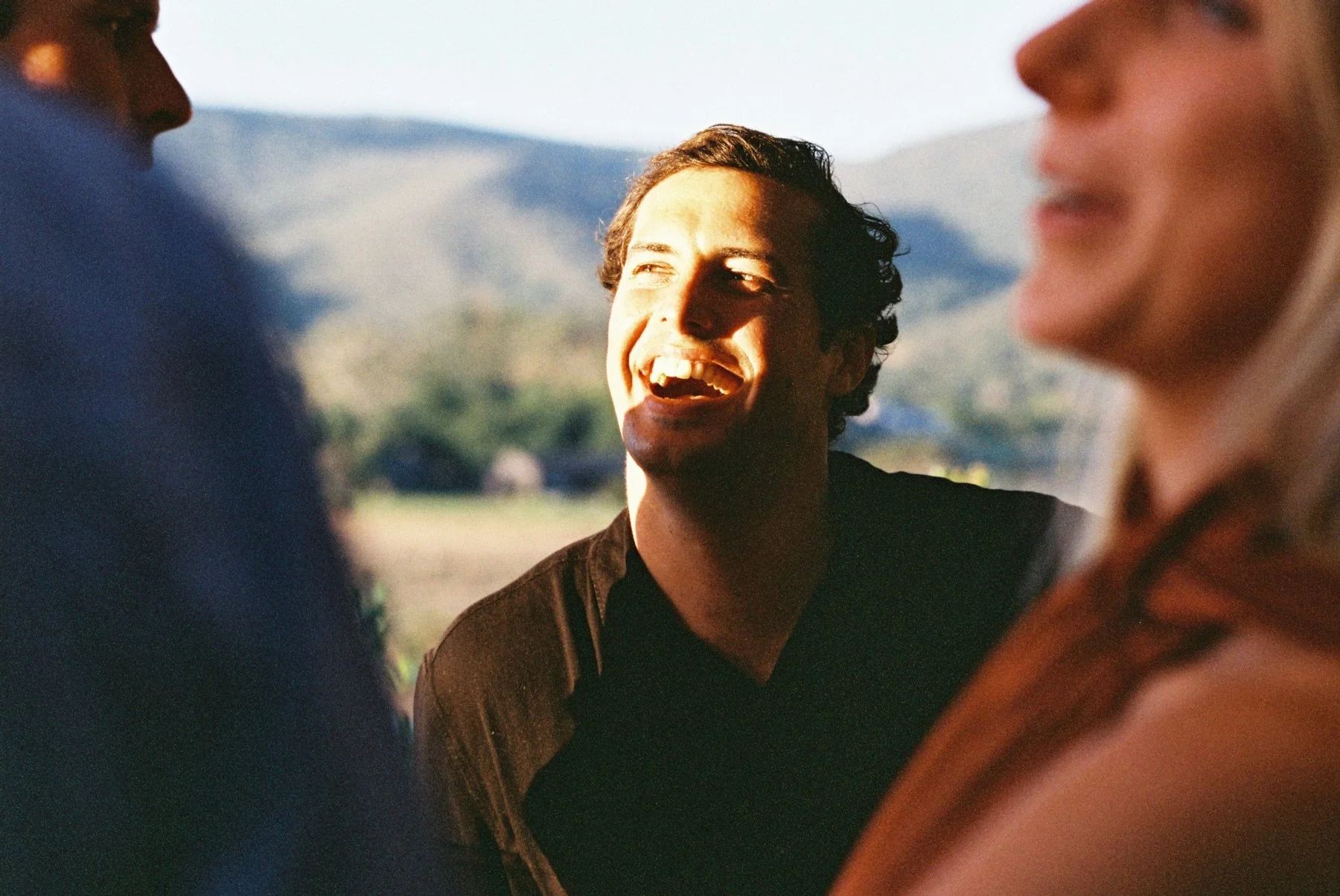 A young man with dark curly hair smiling and laughing, surrounded by blurred faces of other people in an outdoor setting with mountains in the background.