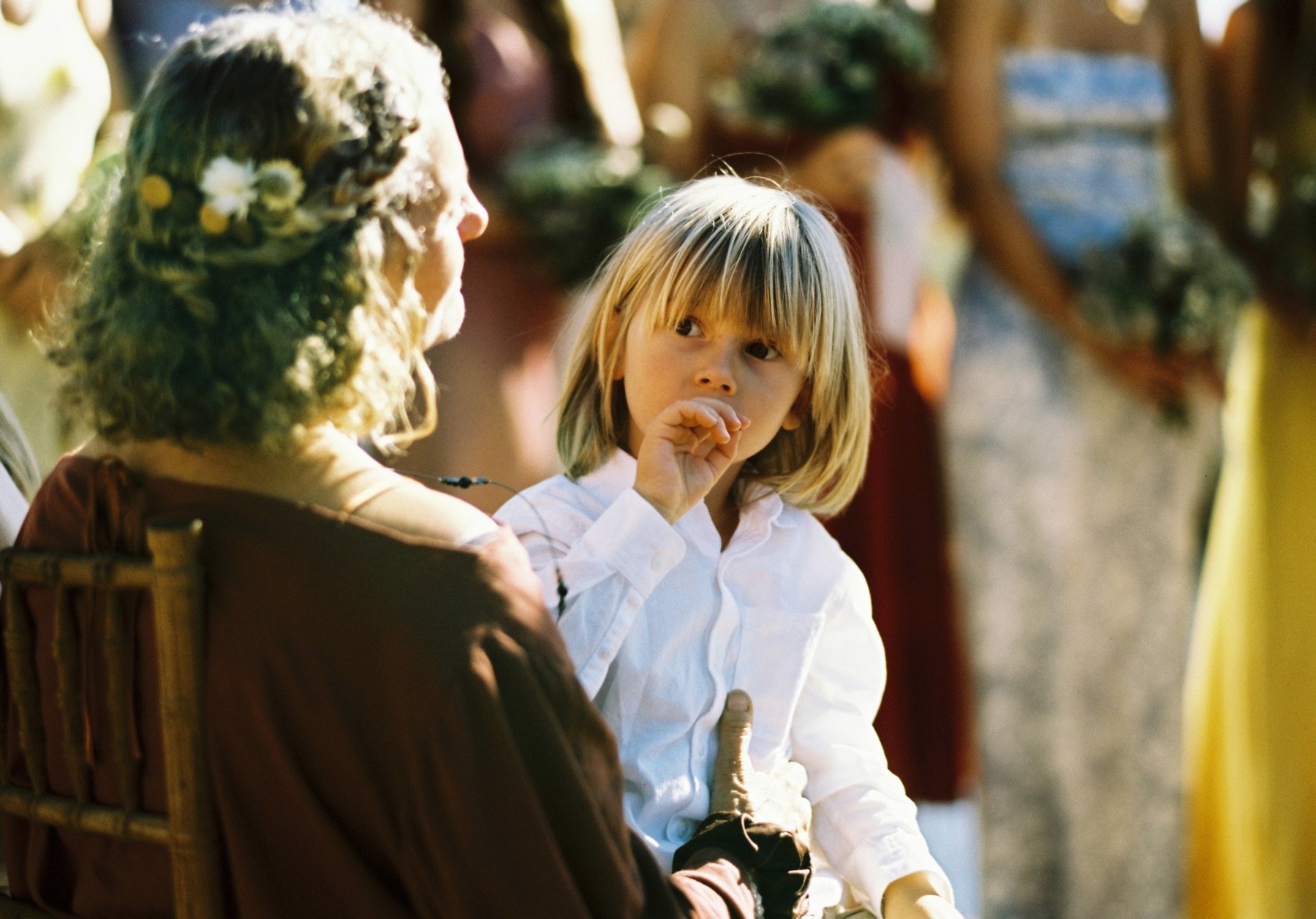 A young boy with blond hair and a white shirt sitting in an outdoor setting, looking at and listening to a woman with curly hair decorated with flowers, who is seated nearby and facing him.