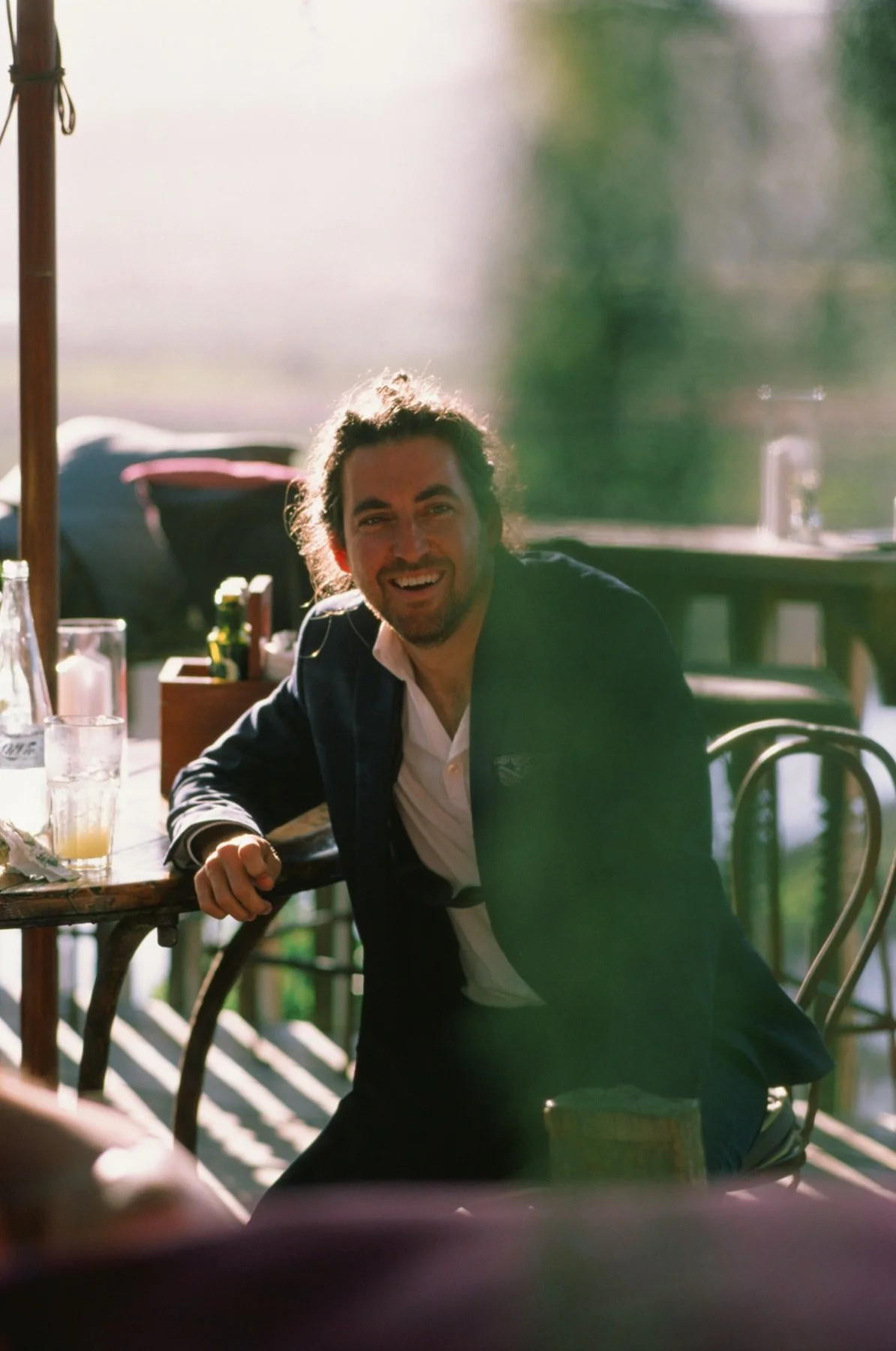 A man in a suit smiling and leaning on a table at an outdoor dining area with sunlight.