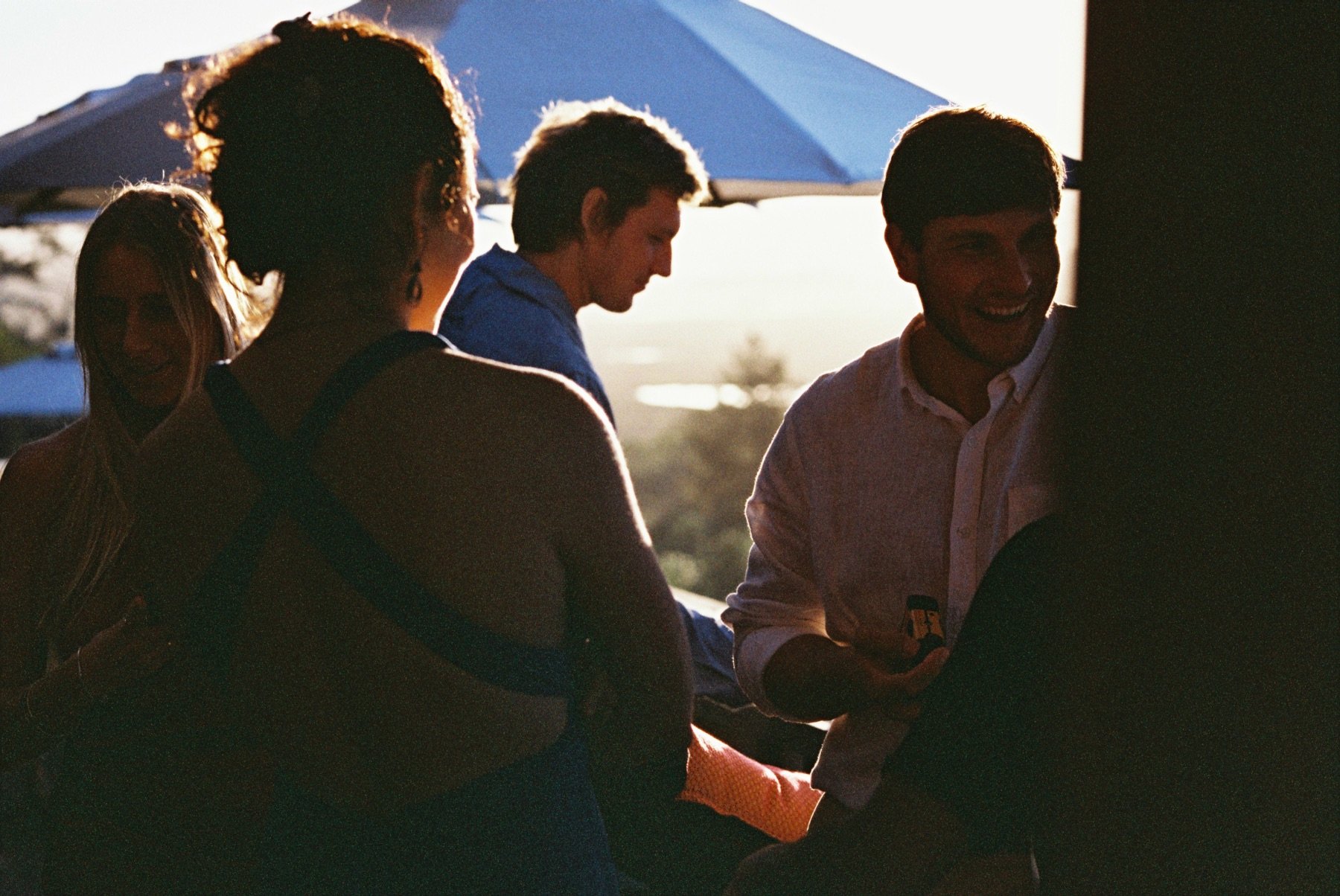 Four people are gathered outdoors near a blue umbrella during sunset. They are engaged in conversation, with some smiling and one person holding a drink. The sunlight creates a backlit scene with shadows highlighting their profiles.