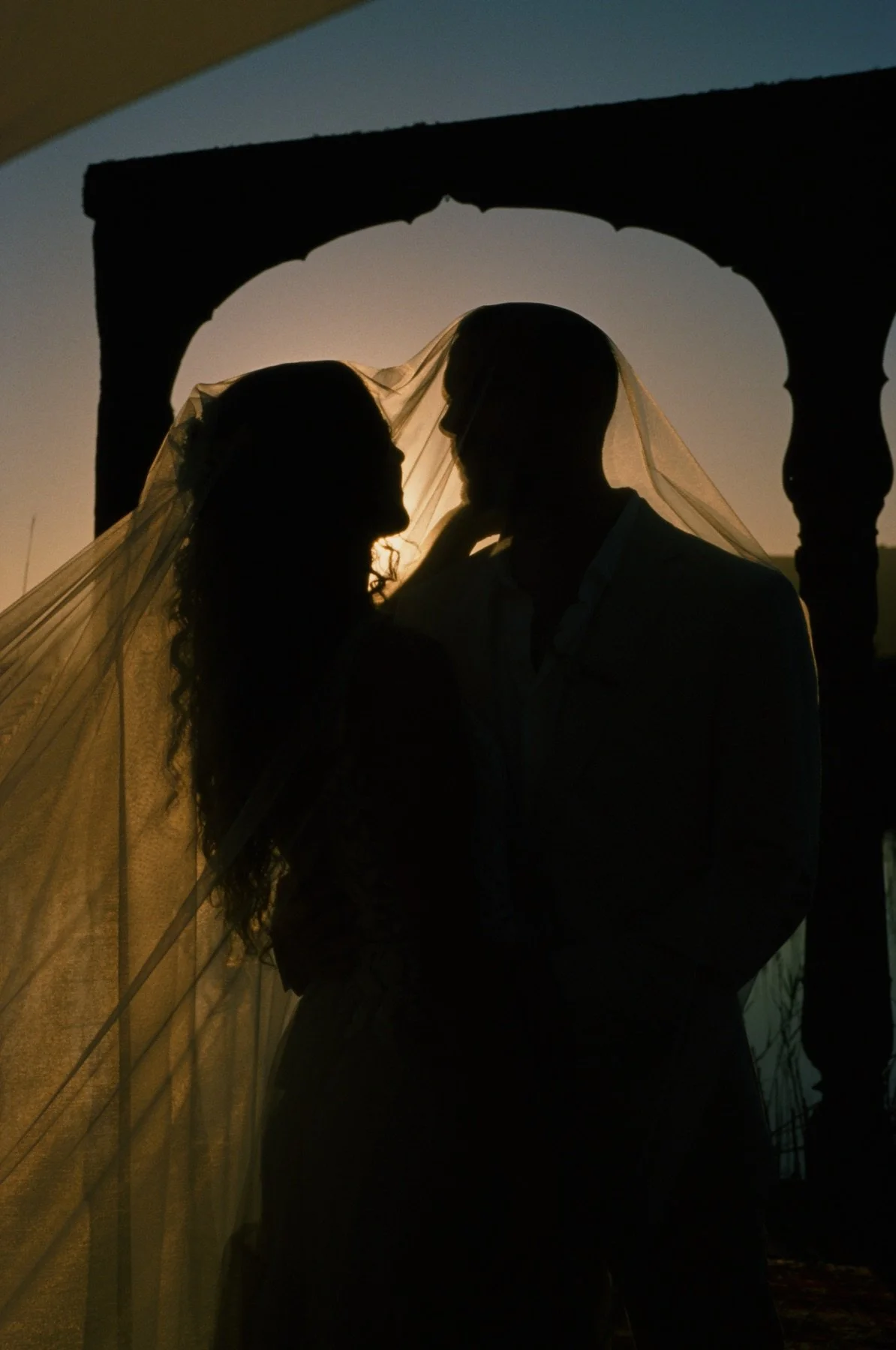 Silhouette of a bride and groom sharing an intimate moment at sunset, with a veil softly flowing and an arch in the background.