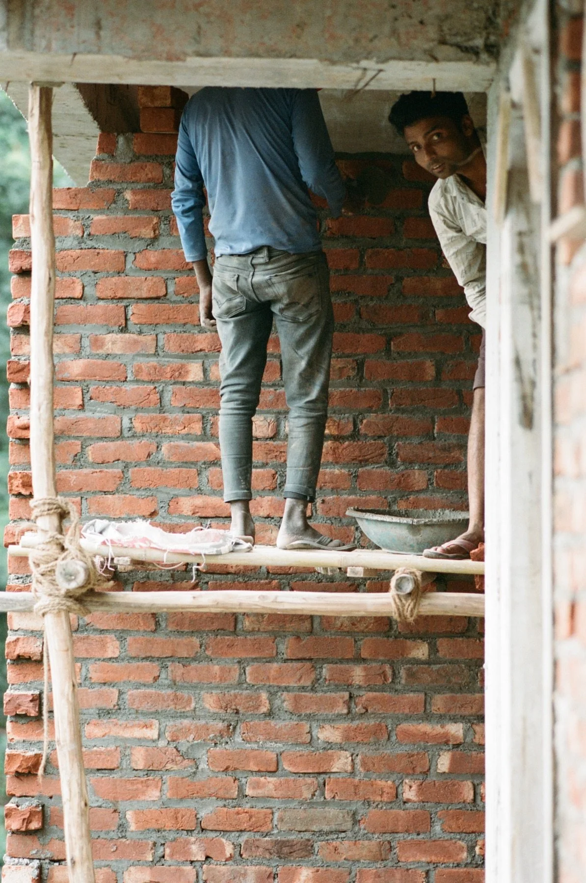 Two people working on a brick wall construction site, with one person facing away and the other peeking from the side, standing on wooden scaffolding.