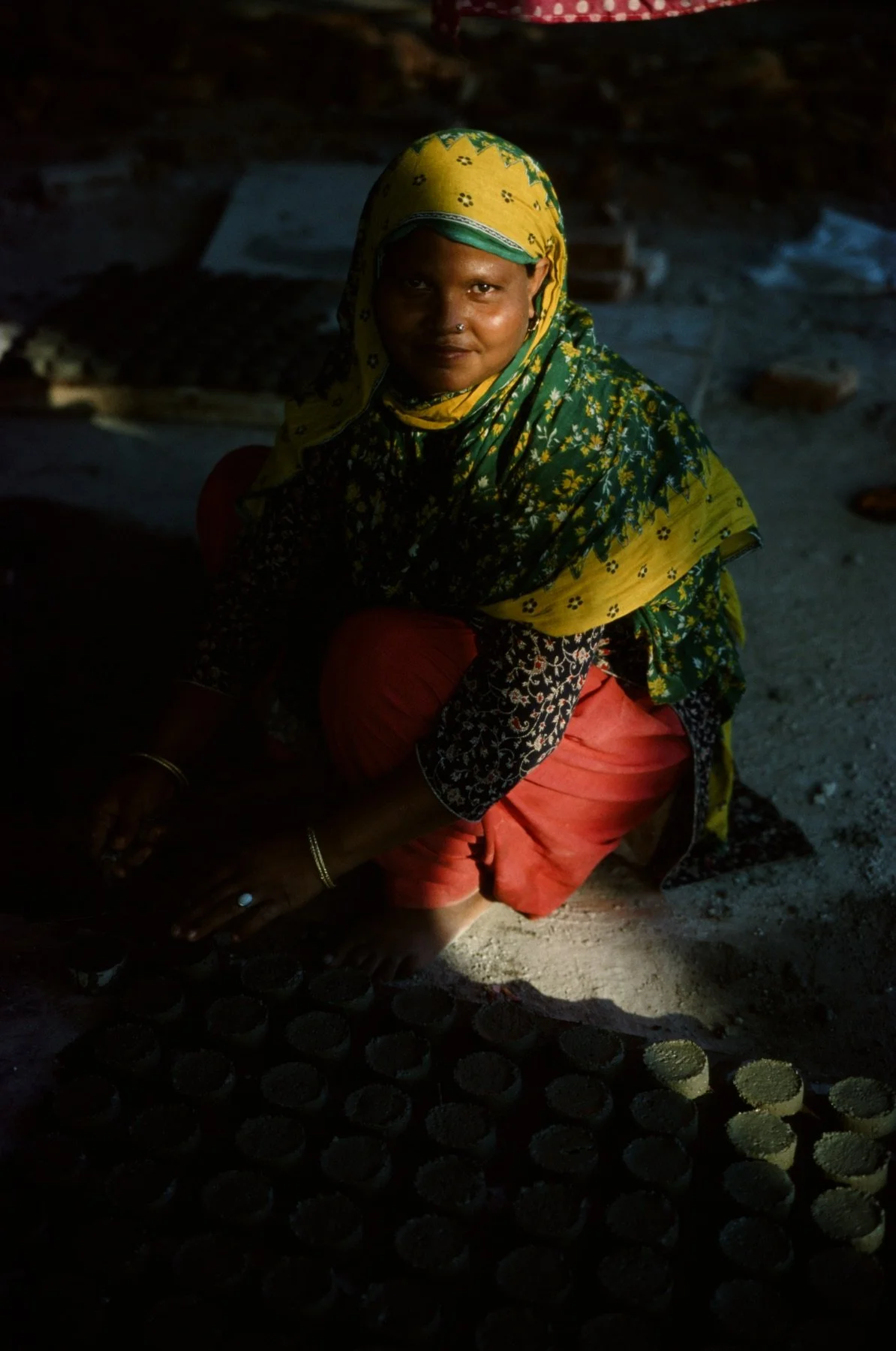 Woman in colorful traditional attire with yellow and green headscarf, sitting on the ground, surrounded by small round objects in a dimly lit setting.