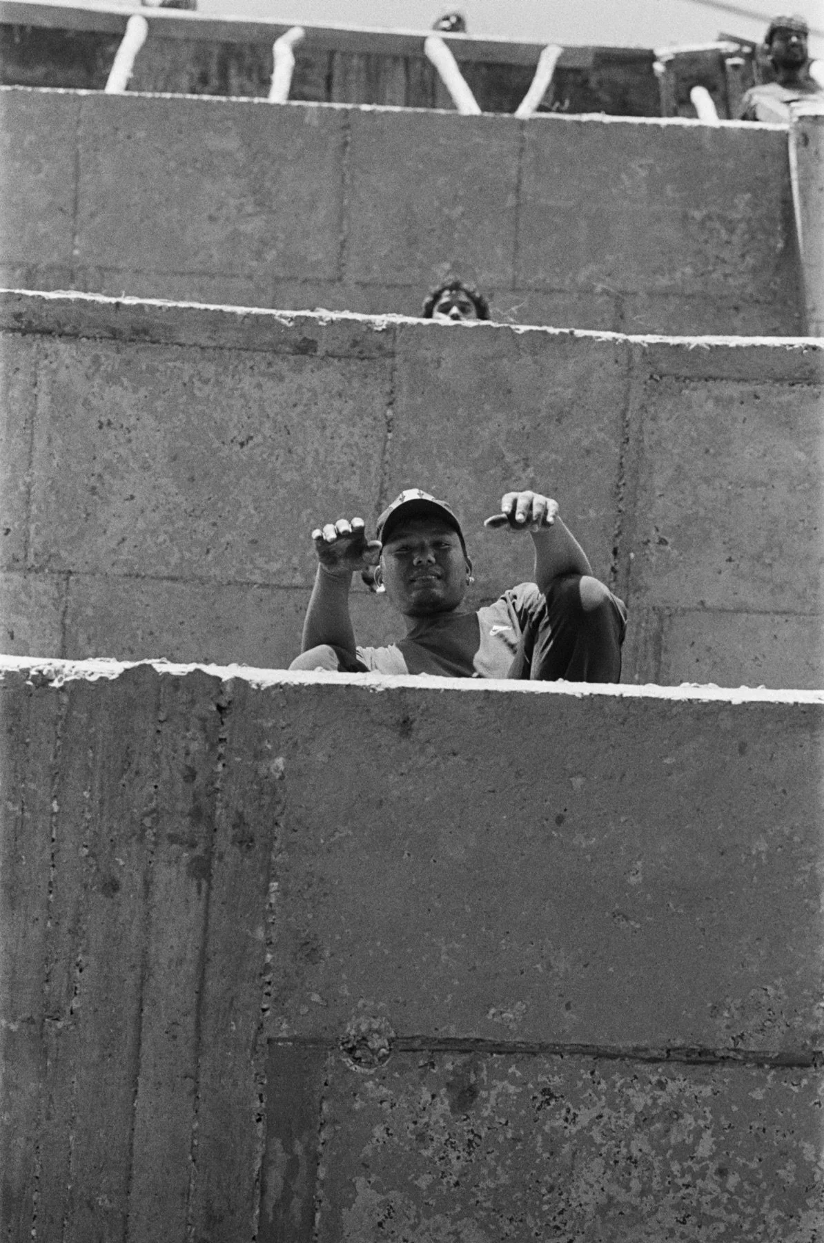 A black and white photo showing three people peeking out from between concrete walls or steps, with a person in the foreground making a gesture with their hands. The person in the foreground appears to be wearing a cap and a casual shirt.
