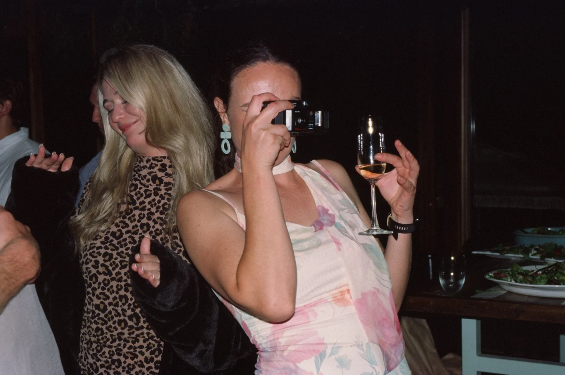 A woman in a floral dress holding a glass of white wine, taking a photo with a camera, at a social gathering or party.