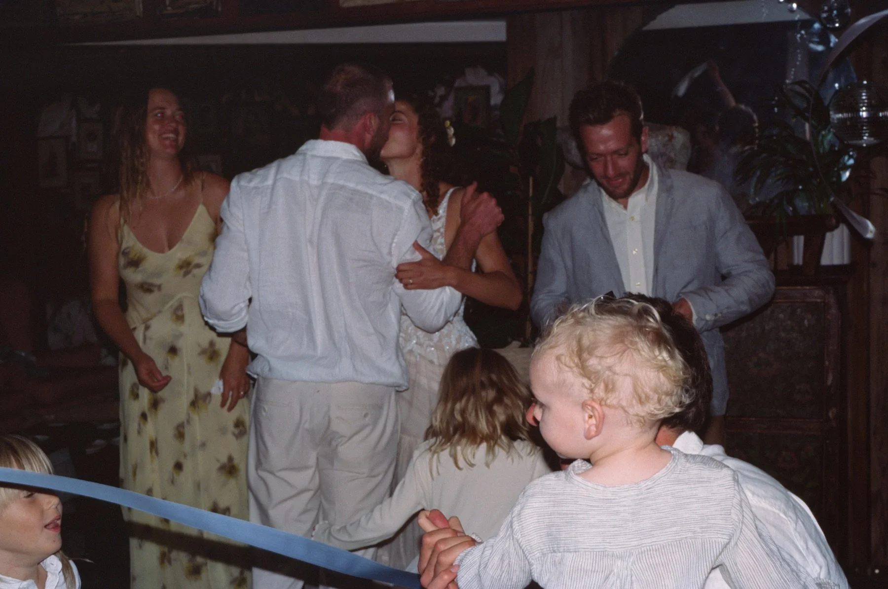 Group of people at a party, with a woman kissing a man on the cheek, while others watch and socialize in a cozy indoor setting.