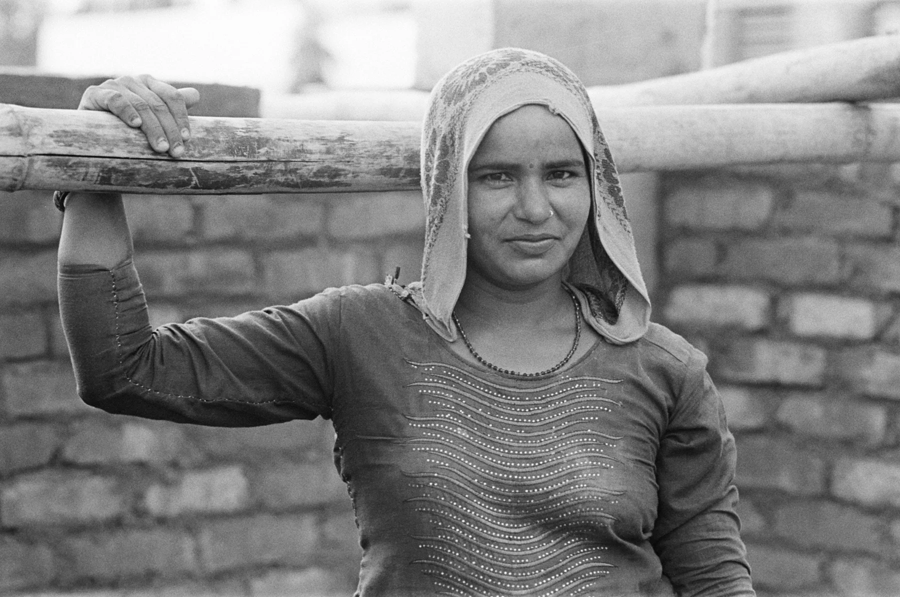 A woman wearing a patterned headscarf and a long-sleeved shirt with decorative stitching, carrying a wooden plank on her shoulder, standing outdoors in front of a brick wall.