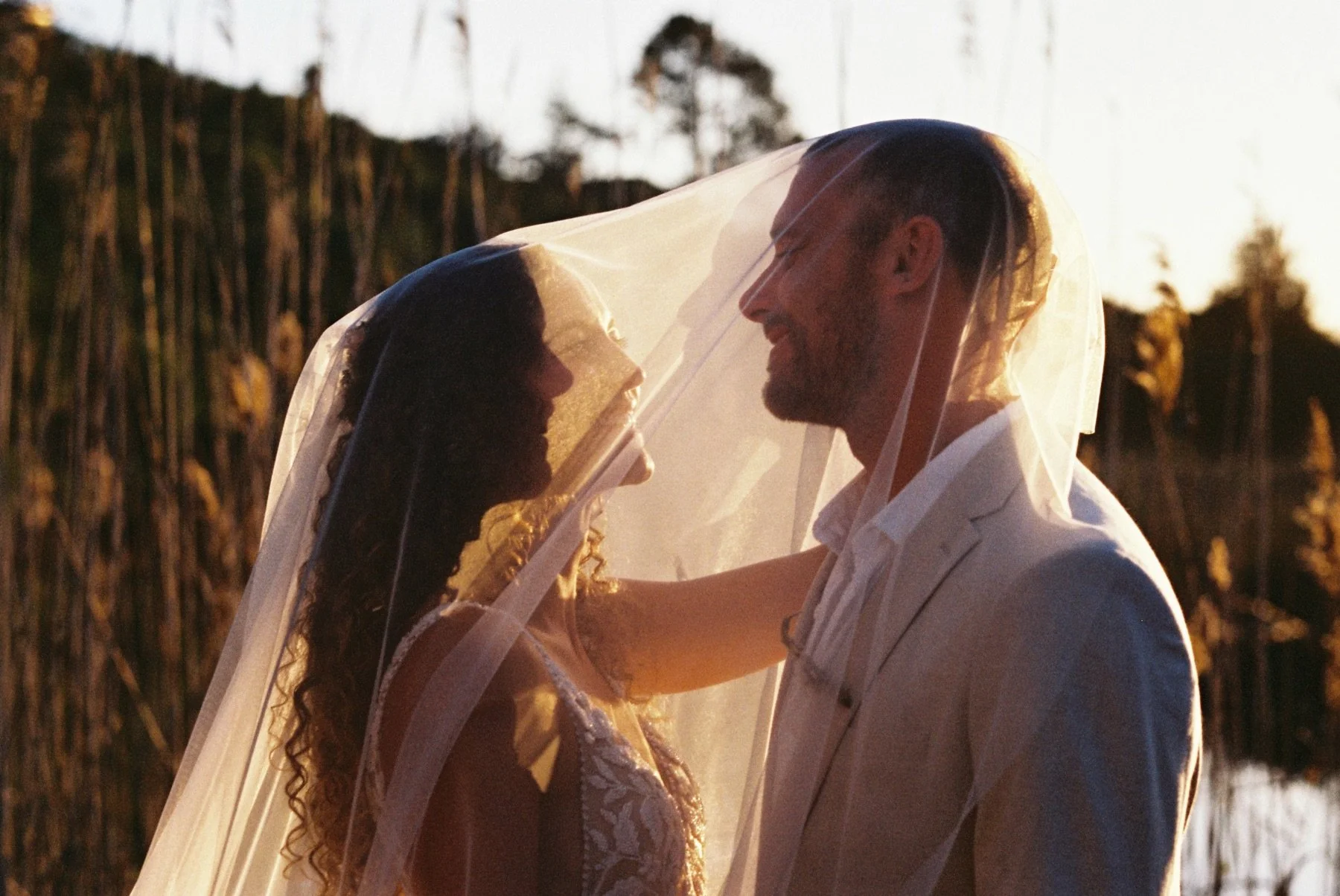 A bride and groom smiling and looking at each other under a veil in an outdoor setting at sunset.