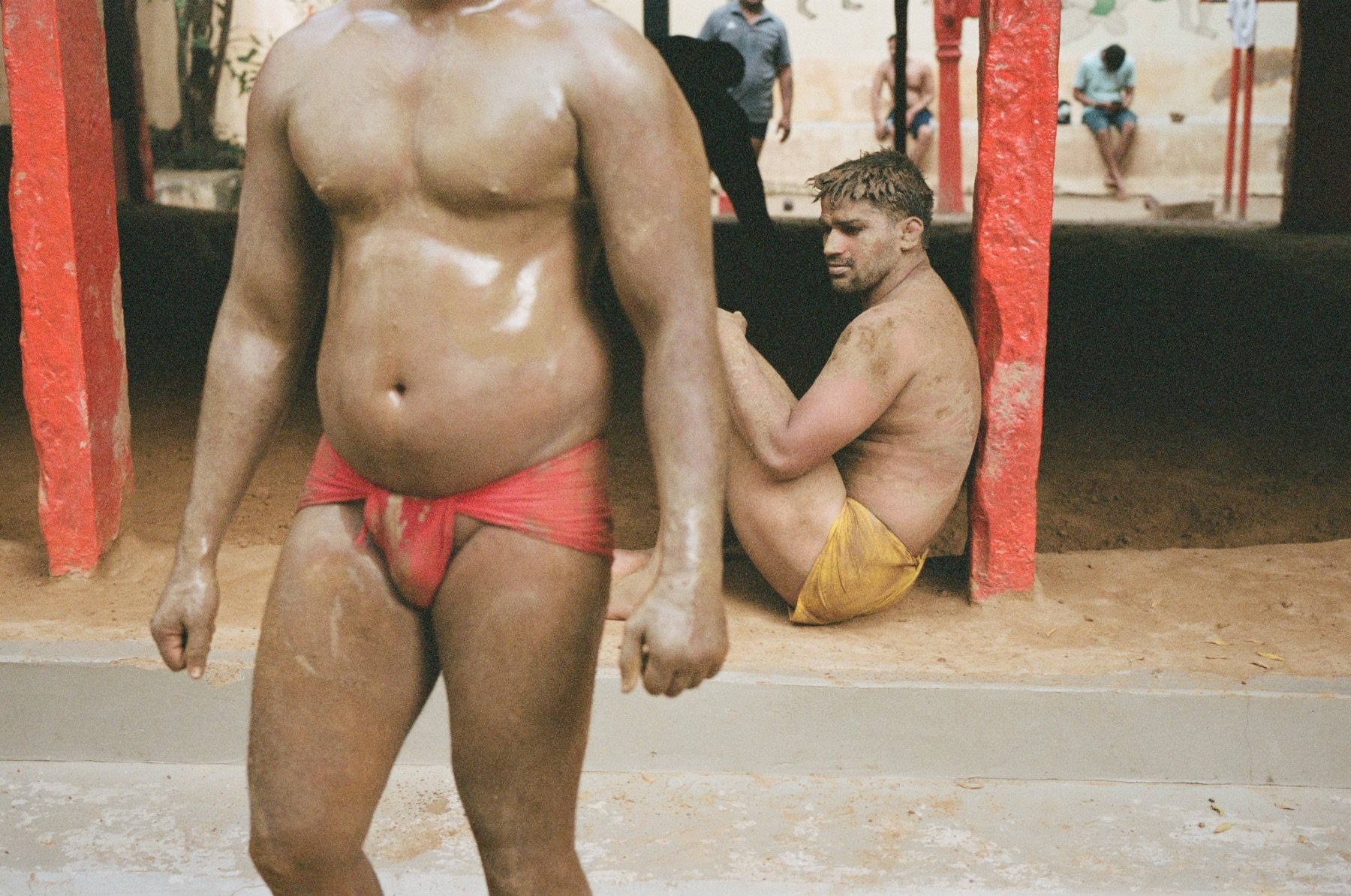Two men in red and yellow swimwear at an outdoor setting, with one seated against a red pillar and the other standing in the foreground.