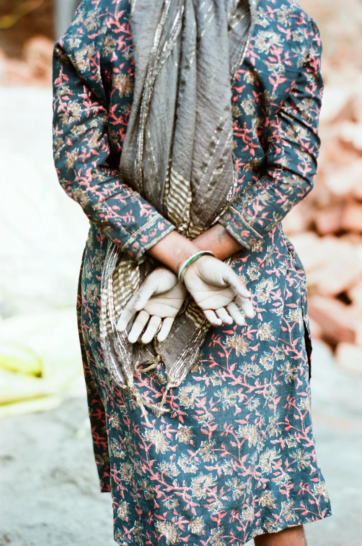 A person wearing a floral dress with a blue background and pink and beige patterns, holding their hands behind their back, with a grey and silver scarf draped over their shoulders.